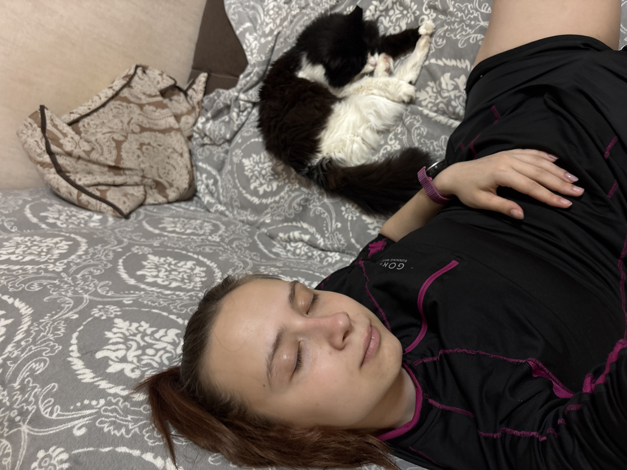 A young woman is lying on a bed with a patterned gray and white duvet cover. Her eyes are closed, and she appears to be sleeping. She wears a black long-sleeved shirt with magenta trim and a pink fitness tracker on her left wrist. To her upper right, a long-haired black and white cat is curled up, also seemingly asleep. A patterned blanket or pillow rests on the bed to the upper left. The location is unspecified and not necessarily related to Прикамский, Russia.