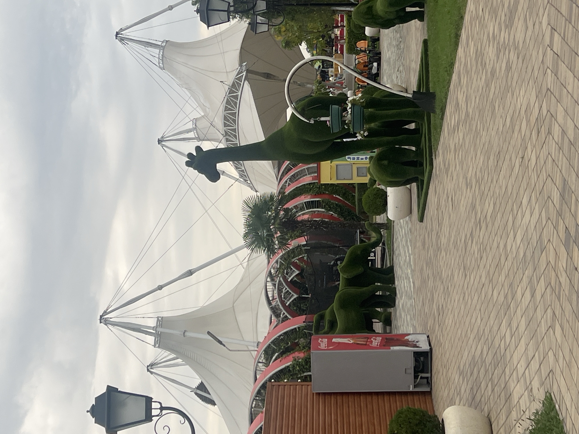 The image captures an outdoor scene featuring topiaries shaped like animals, including elephants and a giraffe, set against a backdrop of large, white, tent-like structures. In the foreground, a paved walkway with a herringbone pattern leads through the area. To the left, a gray refrigerator with a Coca-Cola advertisement is visible next to a wooden wall. The environment appears to be a public space or a park, possibly an amusement park or a landscaped area in the city of Grozny, Russia, given the context provided. The sky is overcast with clouds.