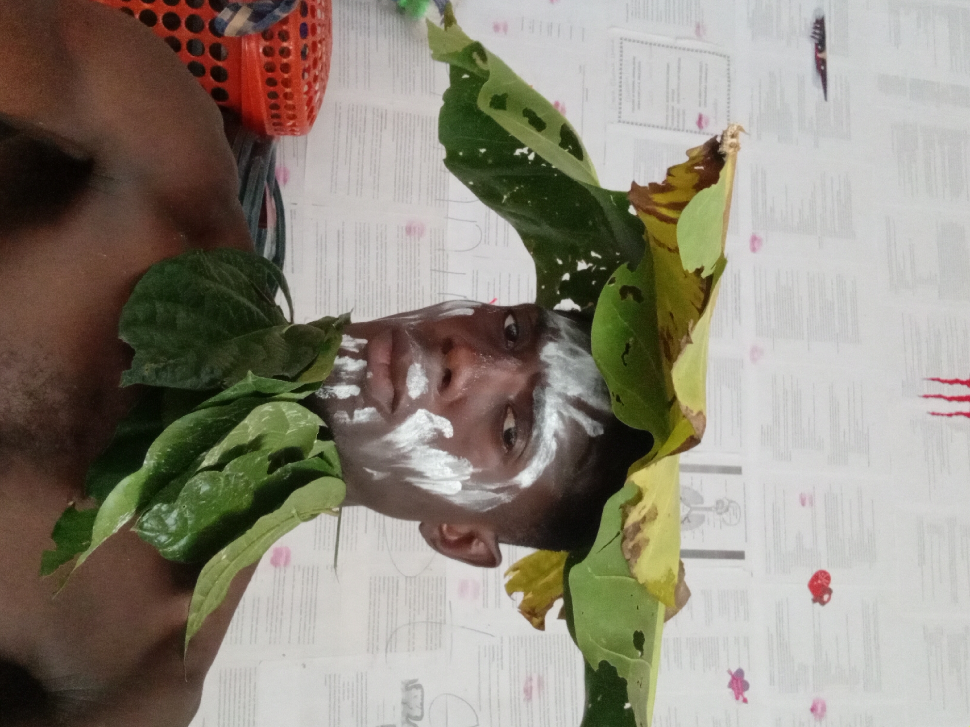 A medium, rotated, close-up shot depicts a dark-skinned man in Akwete, Nigeria, adorned with white face paint and green leaves, posing in front of a wall covered in printed papers.
The man has white paint smeared across his forehead, cheeks, and chin. He is decorated with a leafy collar of small, shiny green leaves on his left side and a makeshift crown of large green and yellow leaves. His left shoulder and upper chest are visible.
Behind him is a white wall covered with printed text, diagrams, and markings. At the top left, a red plastic basket is partially visible. Streaks of red paint or ink are present in the upper right area of the wall. Other small colored markings and drawings are scattered across the wall.