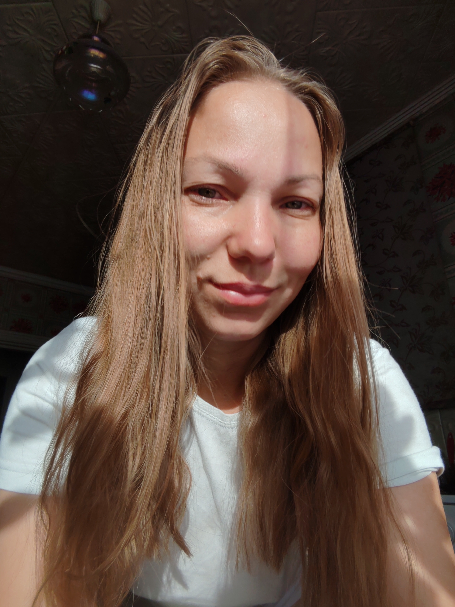 A close-up, selfie-style portrait of a young woman with long, light brown hair, wearing a white t-shirt. She is looking at the camera with a slight smile. The background consists of a patterned ceiling, a dark wall with floral wallpaper, and a ceiling fan. The lighting suggests natural sunlight streaming in from the side. The setting appears to be an indoor residential space. The location is not discernible from the image itself.