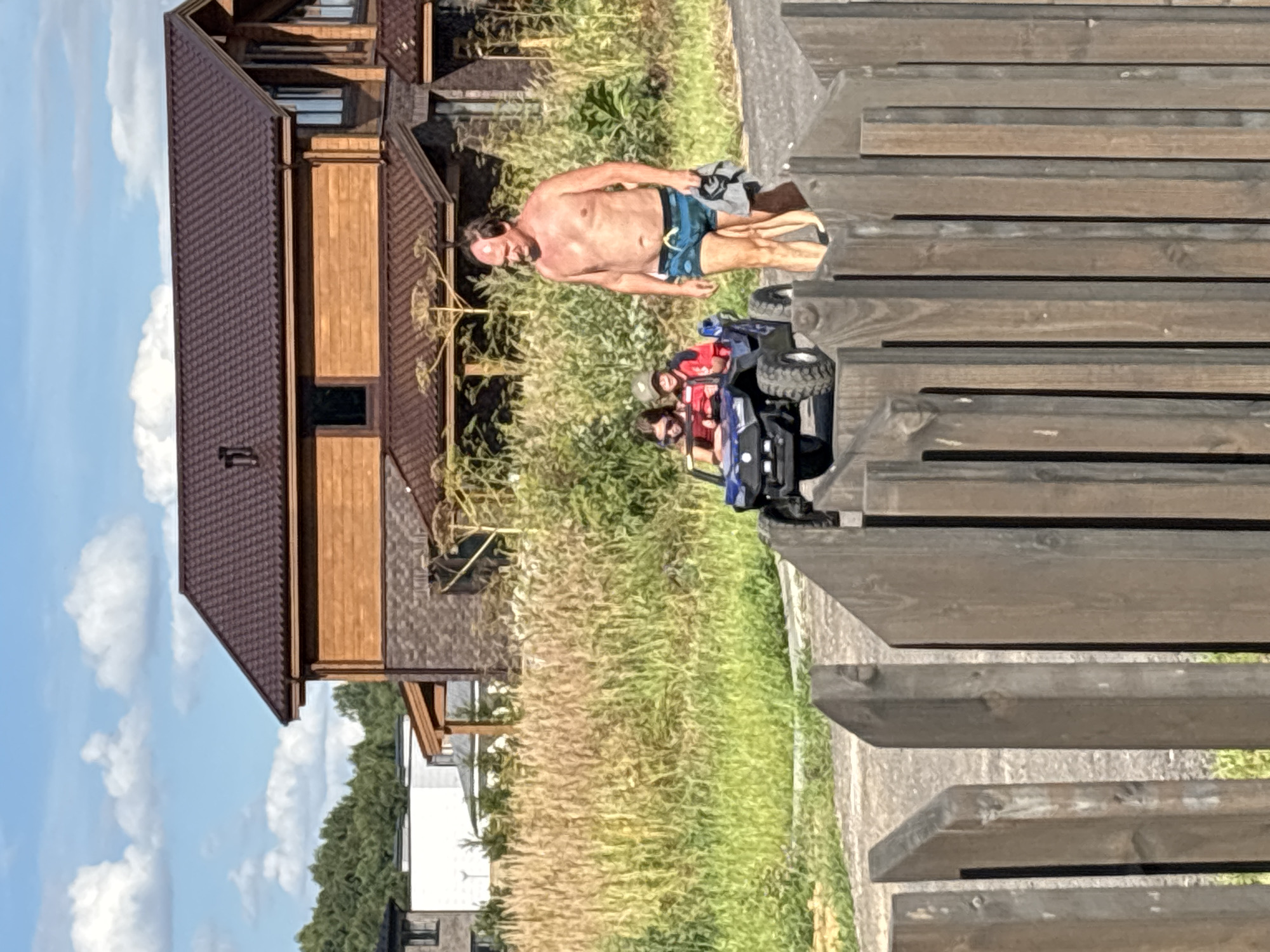 A man, shirtless and wearing blue swimming trunks, walks near a wooden fence in Zvenigorod, Russia. He carries a grey towel in his right hand. In front of him, two children sit in a blue toy car, a girl in a red top in the driver's seat and a boy wearing a green cap behind her. The background features a large wooden house with a dark brown roof, tall dry grass, and green trees under a blue sky with scattered clouds.
