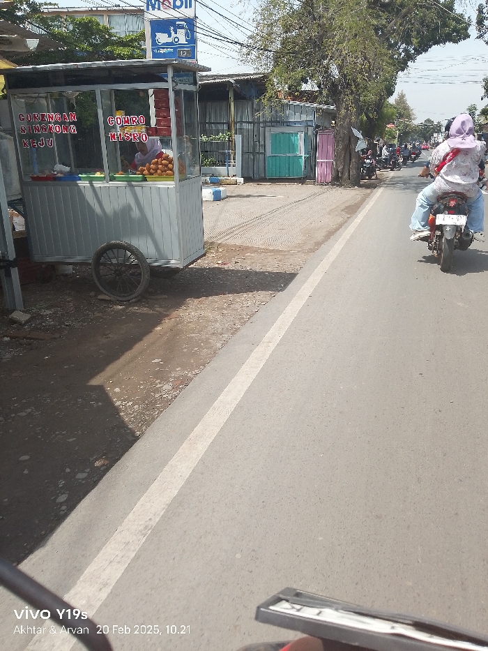 The image depicts a vibrant street scene in Patrol, Indonesia, captured during late morning on a sunny day. In the foreground to the left, a mobile food stall, made of aluminum and glass, stands on the unpaved shoulder of the road. A vendor is visible inside the stall, attending to an array of fried snacks. The stall advertises "GORENGAN SINGKONG KEJU" (Fried Cassava Cheese) and "COMRO MISRO," popular Indonesian street foods.
To the right, an asphalt road with a clear white line stretches into the distance. A person riding a motorcycle, seen from behind, is moving away from the viewer. The rider is dressed in a light-colored patterned shirt, blue jeans, and a purple head covering. Several other motorcycles and vehicles can be observed further down the road.
The background features various structures, including what appear to be simple buildings, some with corrugated metal siding, and a large tree providing shade. A blue and white Mobil gas station sign, partially visible, displays the number "13." The area immediately surrounding the food stall is dirt and gravel.
The bottom left of the image includes a watermark with text: "vivo Y19s Akhtar & Arvan 20 Feb 2025, 10.21," indicating the device, photographer's names, date, and time.