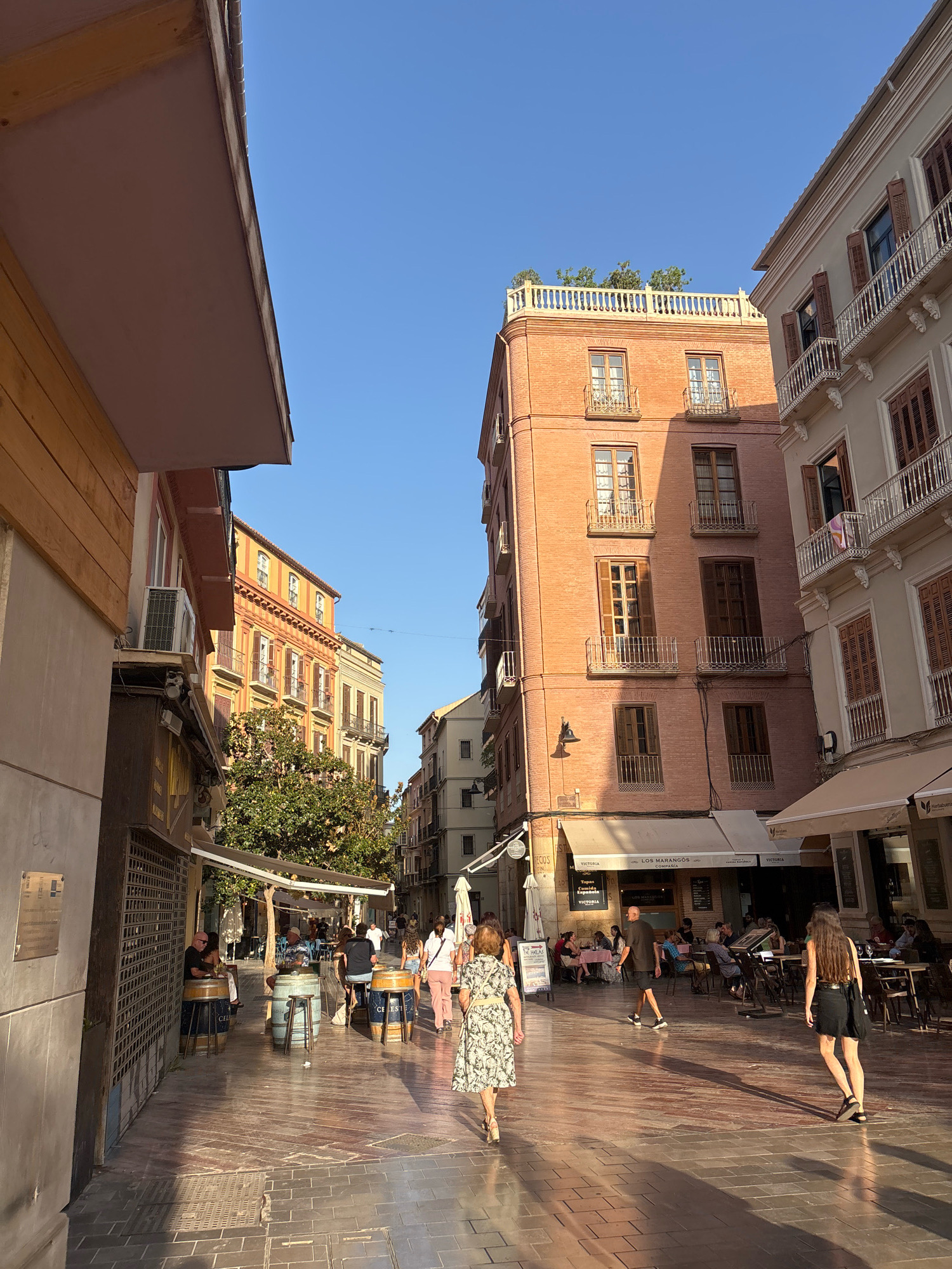 The image depicts a street scene in Malaga, Spain, likely in the city center, with a clear blue sky overhead. The perspective is from ground level, looking down a pedestrianized street lined with buildings. The street is paved with reddish-brown bricks, and in the foreground, a woman in a floral dress walks away from the camera, casting a long shadow. To her right, another woman in a black dress and sandals also walks away, her shadow similarly stretched.
On the left side of the street, a building with wooden siding features an outdoor seating area with tables and chairs, and a large metal grate. Further down, awnings extend over storefronts and more outdoor seating areas are visible, occupied by people. A large tree with green foliage provides shade.
The buildings are multi-story, with varying architectural styles. The building directly ahead and to the right is made of pinkish brick and has several floors with balconies and wooden-shuttered windows. A rooftop garden is visible at the top. To its right, a lighter-colored building exhibits balconies with ornate railings. On the left, a building with yellow and orange facades has multiple windows and balconies. The street is bustling with people, suggesting it's a popular area for dining and strolling. The lighting indicates it's late afternoon or early evening, given the long shadows.
