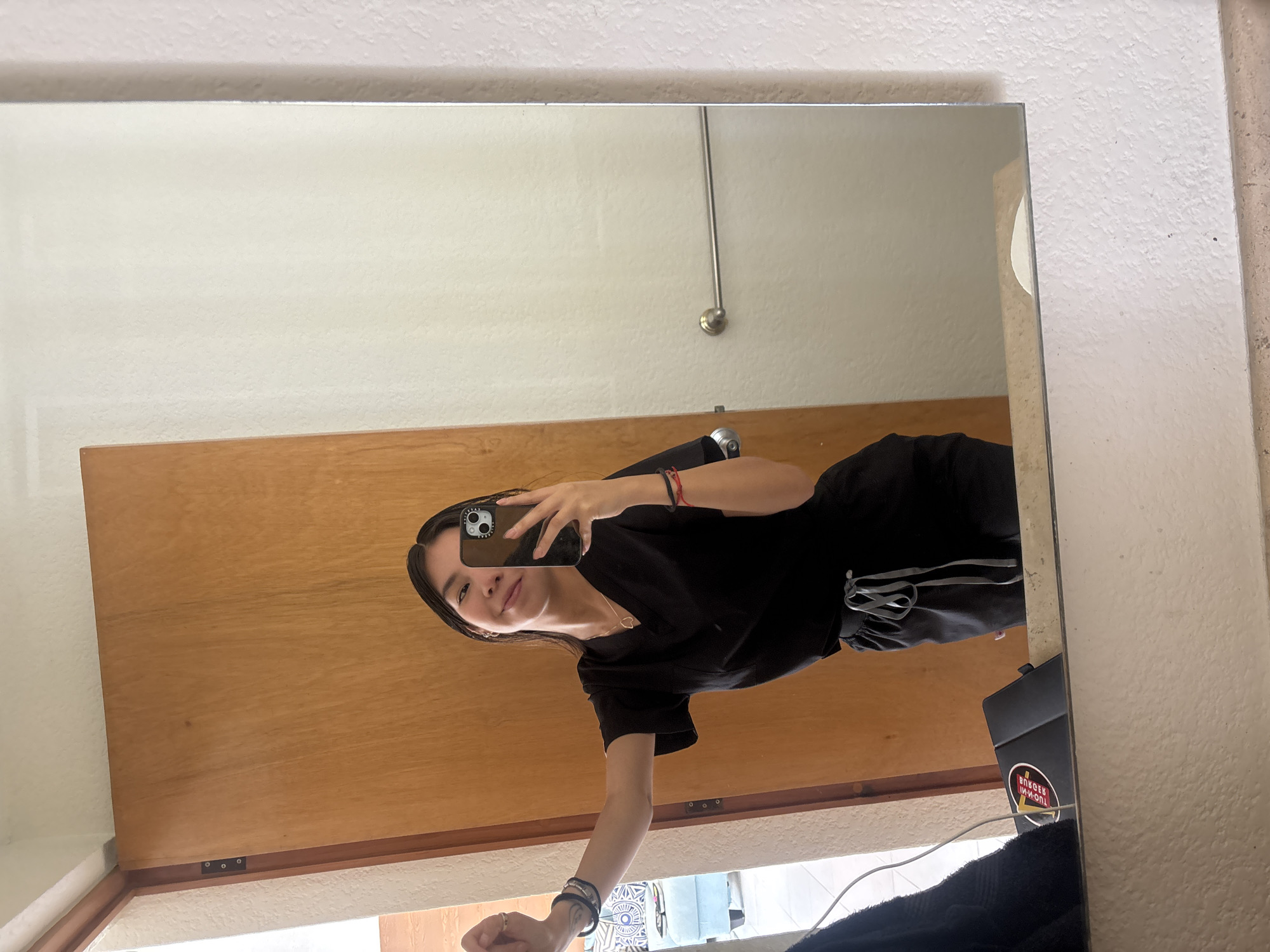 A woman is taking a mirror selfie. She is wearing a black top and black pants with a gray stripe. Her phone is in her left hand. She is standing in front of a door. There is a white wall in the background. A mirror is visible, showing a reflection of the woman and the surrounding environment. The image does not contain sufficient details to determine a location in Puebla, Mexico.