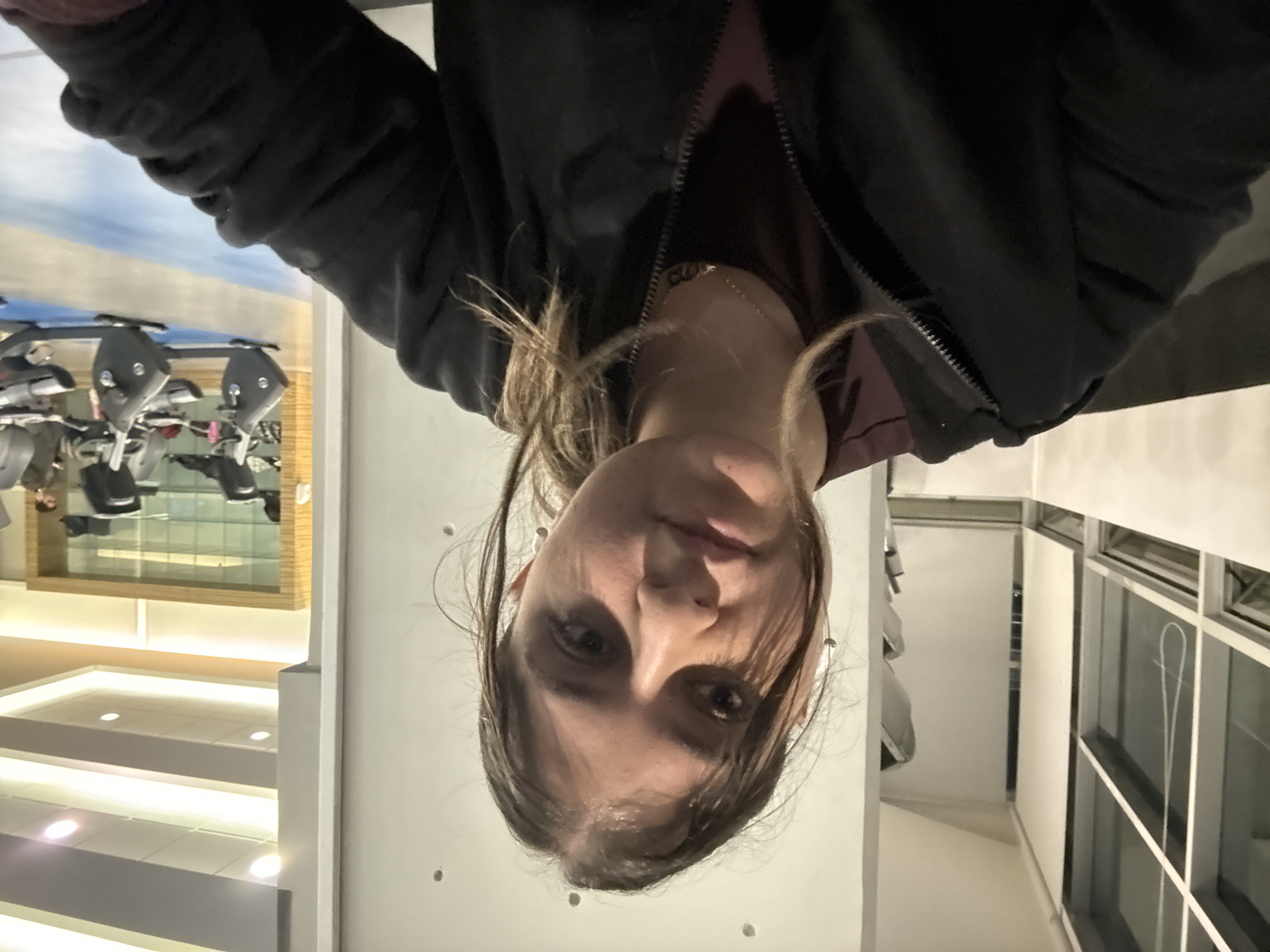 A selfie-style photograph, originally rotated, captures a young woman looking directly into the camera from a downward angle. She has brown hair, which falls around her face, and is wearing a dark jacket, possibly black, over a maroon or dark red top, with a visible zipper. Her expression is neutral.
The background suggests an indoor setting, likely a fitness center or a public building with gym facilities. To her left, reflected in a large mirror, are multiple pieces of exercise equipment, appearing to be stationary bikes or similar cardio machines. Above this reflected gym area, the ceiling features a large panel designed to resemble a light blue, cloudy sky. Below the gym reflection, there are brightly illuminated white steps or shelves, suggesting a modern architectural element or display area. To her right, a plain white wall and a window with white frames are visible. The lighting appears to be artificial, typical of an indoor environment, making the specific time of day unclear. There is no visible text in the image.