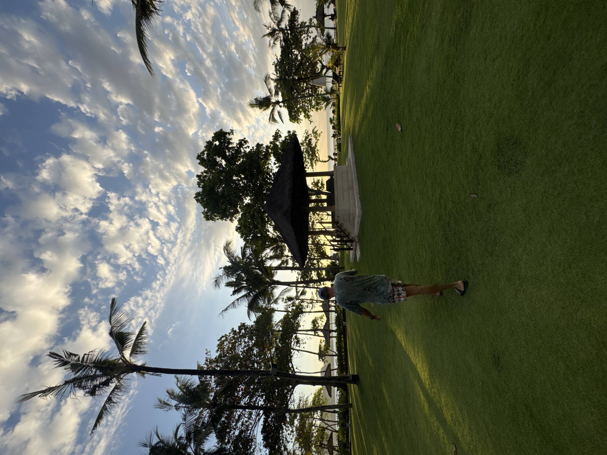 The image shows a man walking on green grass in a tropical location in Kuta Selatan, Indonesia. Palm trees line the edge of the grass near a beach. A small open structure with a thatched roof is also present. The sky is partly cloudy with streaks of light. The angle is rotated slightly, with the sky on the left side of the frame.
