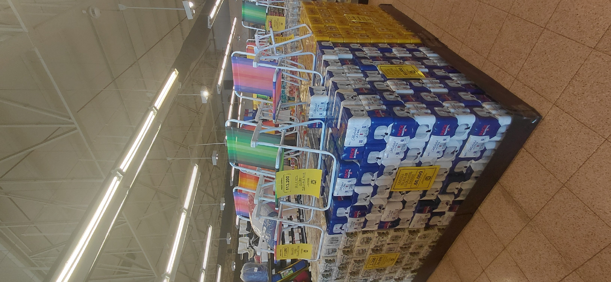 The image shows an interior view of a supermarket in Encarnación, Paraguay. Displayed prominently are several colorful beach chairs with rainbow-striped fabric. These chairs are set up on top of stacked pallets of goods. Below the chairs, and forming the base of the display, are numerous blue and white packages, which appear to be multi-packs of toilet paper. Some of these packages have yellow price tags that read "113.200" and "60.000". To the right of the toilet paper stacks, there are yellow stacks of what appear to be beverage cans, also with some yellow signage. The ceiling features industrial lighting and exposed support structures. The floor is tiled in a beige and brown speckled pattern. The overall impression is of a retail environment promoting outdoor leisure items alongside household necessities and beverages.