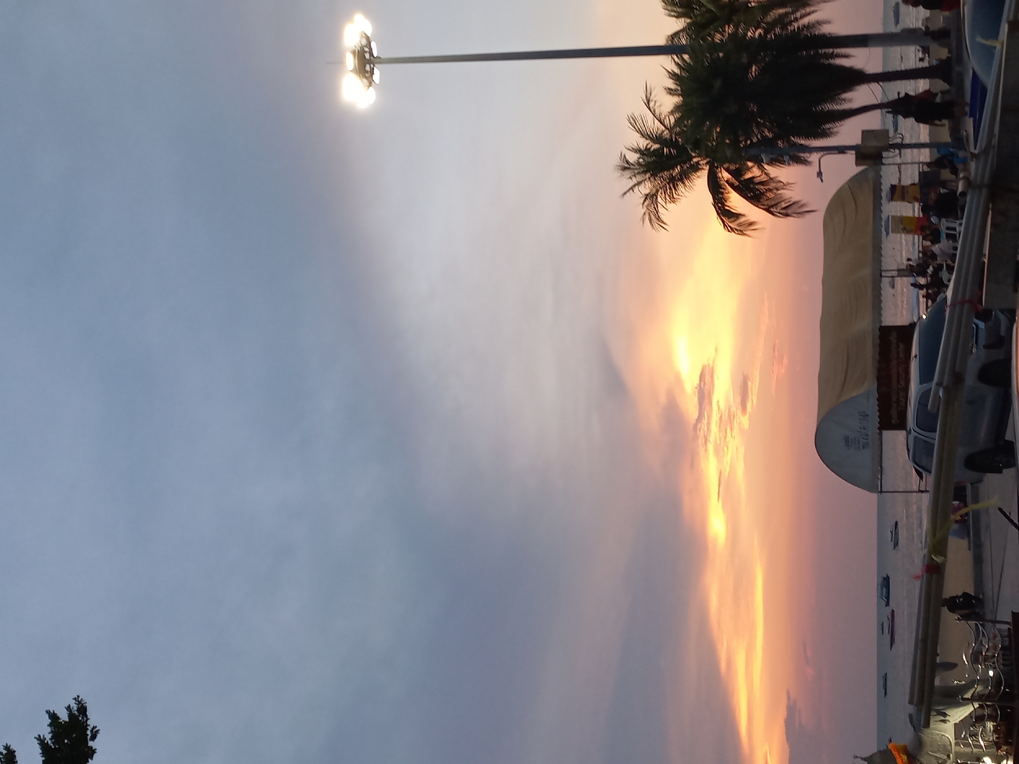 The image captures a picturesque sunset over a coastal area, specifically Pattaya City, Thailand. The sky displays a striking gradient, transitioning from vibrant orange and yellow hues on the right, indicative of the setting sun, to a soft greyish-blue on the left. Patches of clouds are illuminated by the warm light.
A prominent feature in the upper left is a tall pole-mounted streetlight or floodlight, equipped with multiple bright fixtures that emit a strong white light. Below this, several palm trees are silhouetted against the colorful sky on the right side of the frame, adding to the tropical ambiance.
Below the horizon line, a calm body of water, likely the sea, stretches across the scene, reflecting the twilight. Numerous small boats are visible on the water, some appearing to be at anchor or near the shore. Along the right edge of the image, a waterfront area features various structures, including a cream-colored tent or awning with visible Thai script, and parts of vehicles such as cars or motorcycles. Silhouettes of people can be seen in the background, suggesting human activity or leisure by the water.
The scene depicts the transition from day to night, with the artificial lights beginning to glow against the last vestiges of daylight, creating a serene yet lively atmosphere typical of a resort town at dusk.