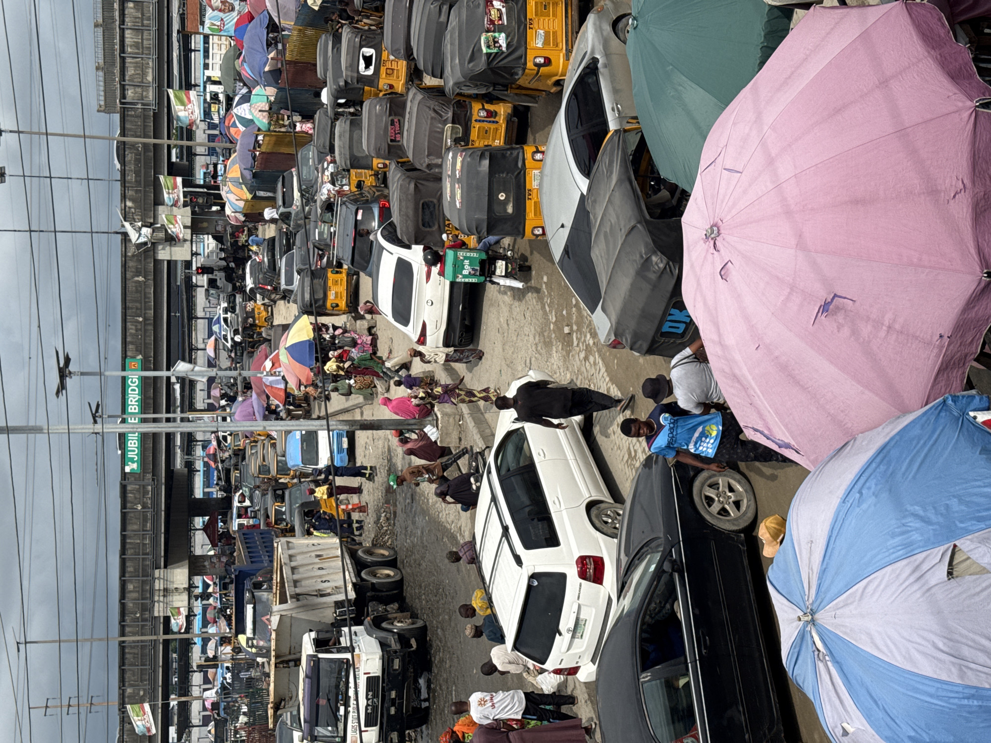 A high-angle, slightly tilted, medium shot captures a crowded street scene in Lekki, Nigeria, under an overcast sky. The foreground is dominated by several large, open umbrellas, a pink one on the right and a blue and white striped one on the left, partially obscuring vehicles and people below.
In the midground, a white SUV is parked, its doors open. Several people are milling around, some appearing to be vendors or shoppers. A blue and white delivery motorcycle, with a green bag emblazoned with "Bolt" on the back, is positioned near the SUV. A line of yellow auto-rickshaws, known as 'Keke Marwa' or 'Keke napep', is also visible, some of them occupied. A black sedan is parked in the lower left, and another silver sedan is partially visible.
The background reveals a busy market or transport hub. A large, white tipper truck with "SPWC" written on its side is prominent, along with other vehicles and more auto-rickshaws. A concrete overpass or bridge, marked with a green sign that reads "JUBILE BRIDGI", stretches across the upper part of the image, with several flags attached to it. Power lines and street lights are also visible overhead. The ground appears to be a mix of paved and unpaved surfaces, with some puddles of water suggesting recent rain. The overall impression is one of a vibrant, bustling urban environment with significant traffic congestion and commercial activity.