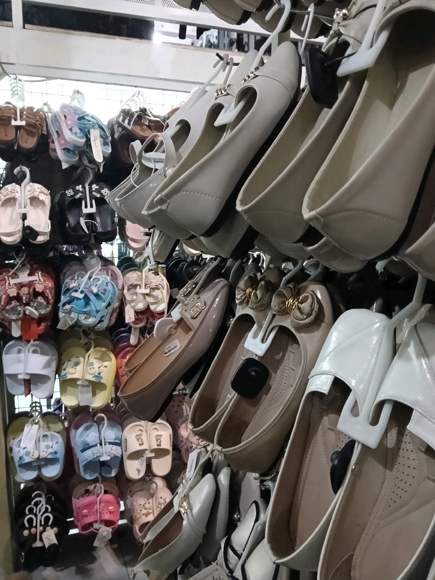 The image shows a retail display of women's shoes and sandals in Dengok, Indonesia. The shoes are arranged on shelves and hanging displays, with a variety of styles and colors. Many of the shoes are flat, ballet-style flats, while others are sandals and flip-flops. Some of the shoes have decorative elements such as bows, pearls, and buckles. The display appears to be in a store or market setting, with a metal shelving unit in the background. The lighting is artificial, suggesting an indoor environment. There are no people visible in the image, and the focus is solely on the merchandise.