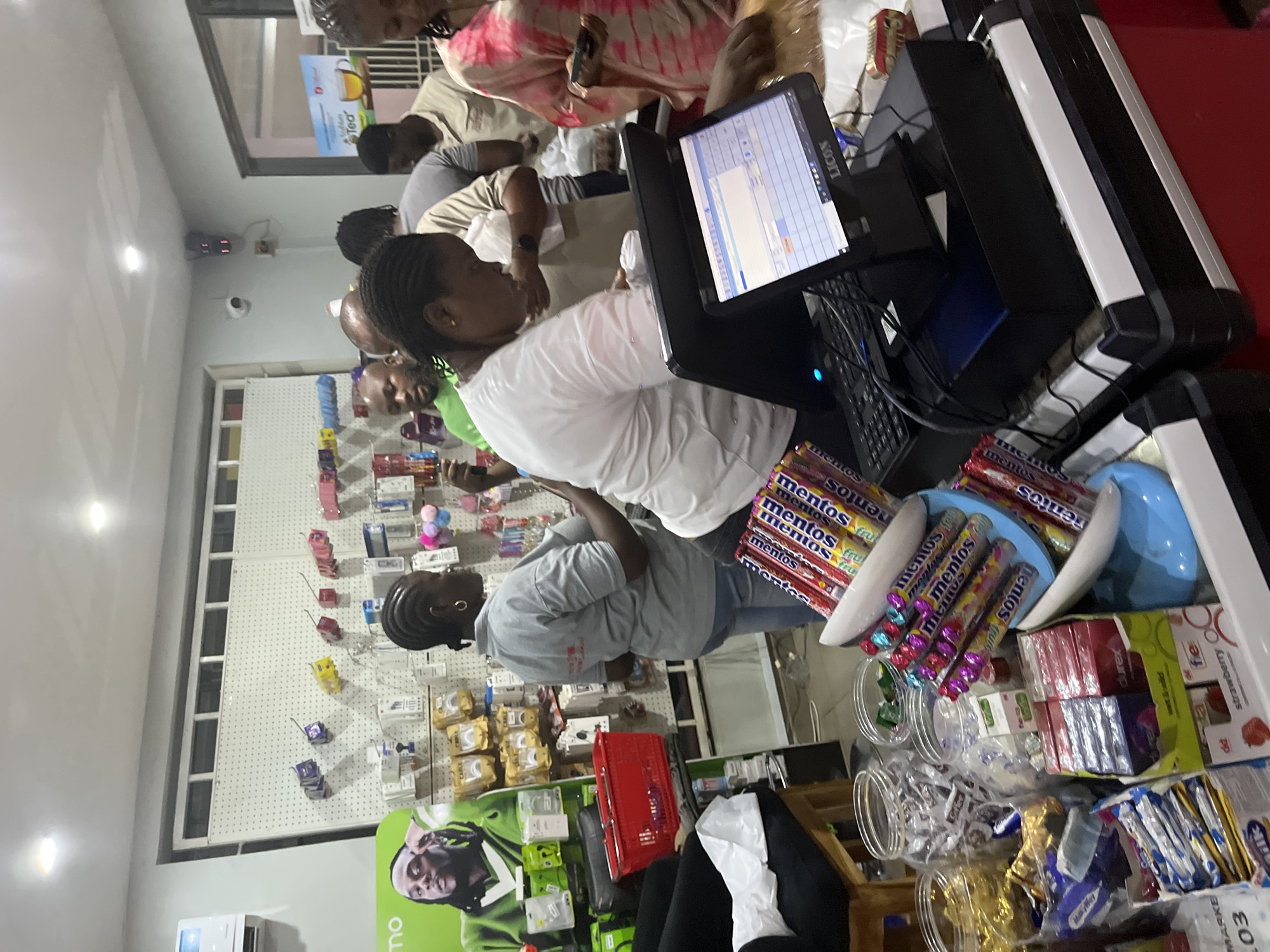 The image depicts a busy checkout counter in what appears to be a retail store or pharmacy in Abuja, Nigeria. Several customers are lined up waiting to make their purchases. A cashier wearing a white shirt is operating the point-of-sale system. The counter is cluttered with various impulse purchase items such as Mentos rolls, candies, condoms, and other small goods. A pegboard display of merchandise can be seen in the background. There is a security camera on the ceiling and a cooler advertising tea is visible near the front of the line. The checkout area is brightly lit.