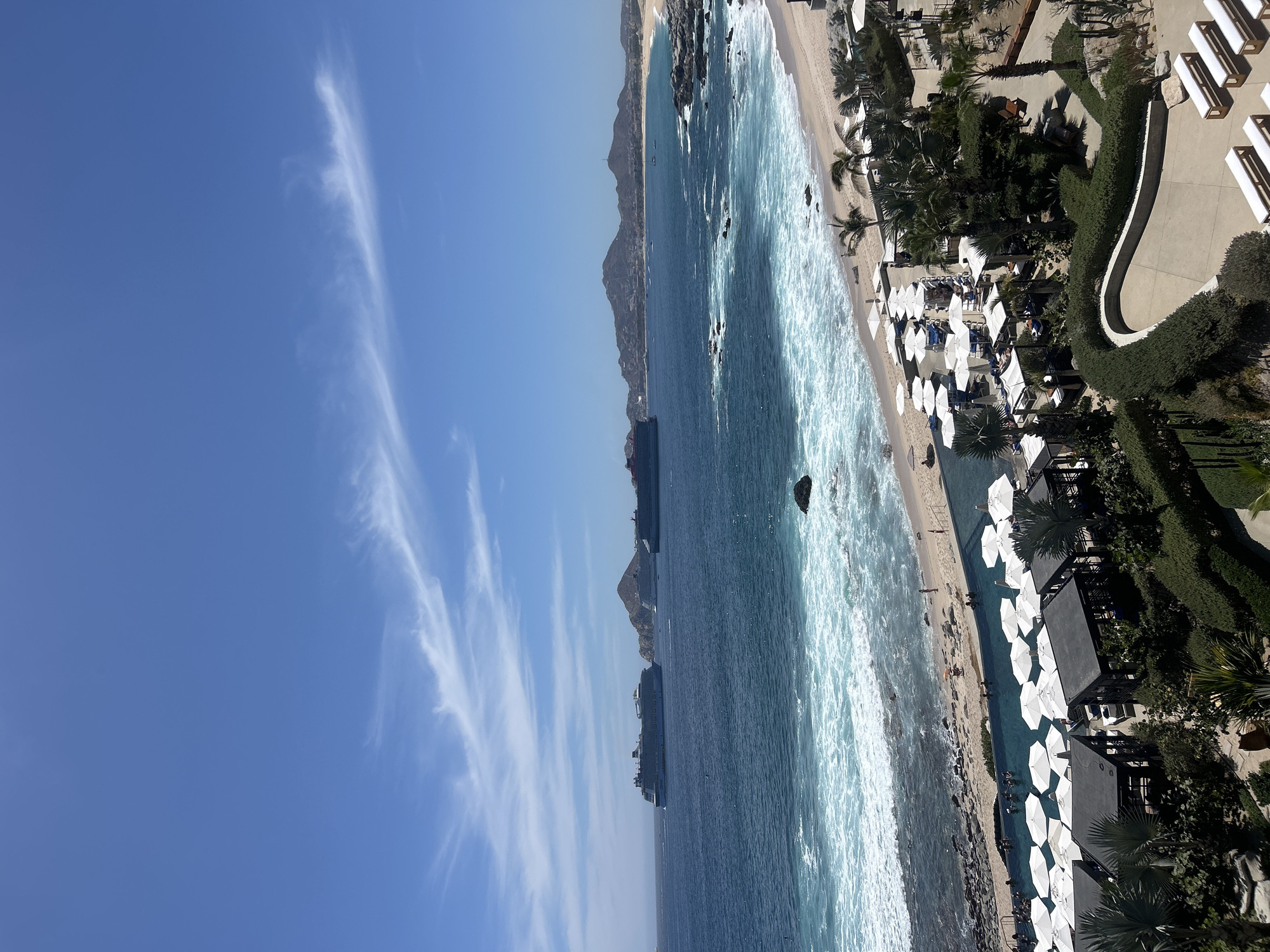 The image shows a beach in Cabo San Lucas, Mexico, with a blue sky containing several wispy clouds. The ocean is turquoise, and the waves are visible. The beach has white sand, and there are people on it. A line of white beach umbrellas runs along the coast. There is a resort with dark green foliage between the beach and a concrete walkway. Two cruise ships are visible on the sea. Mountains are in the distance.
