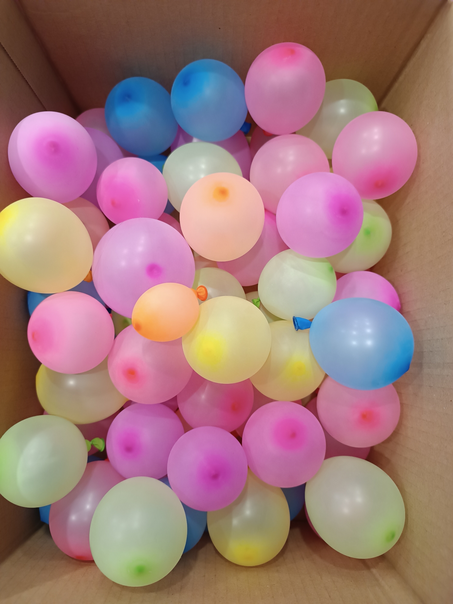 A cardboard box is filled with numerous inflated, small, round balloons in assorted pastel colors including pink, blue, yellow, green, and orange. The balloons are densely packed within the box. The interior of the cardboard box is visible, revealing its brown color. The location of the photograph is not discernible from the image.