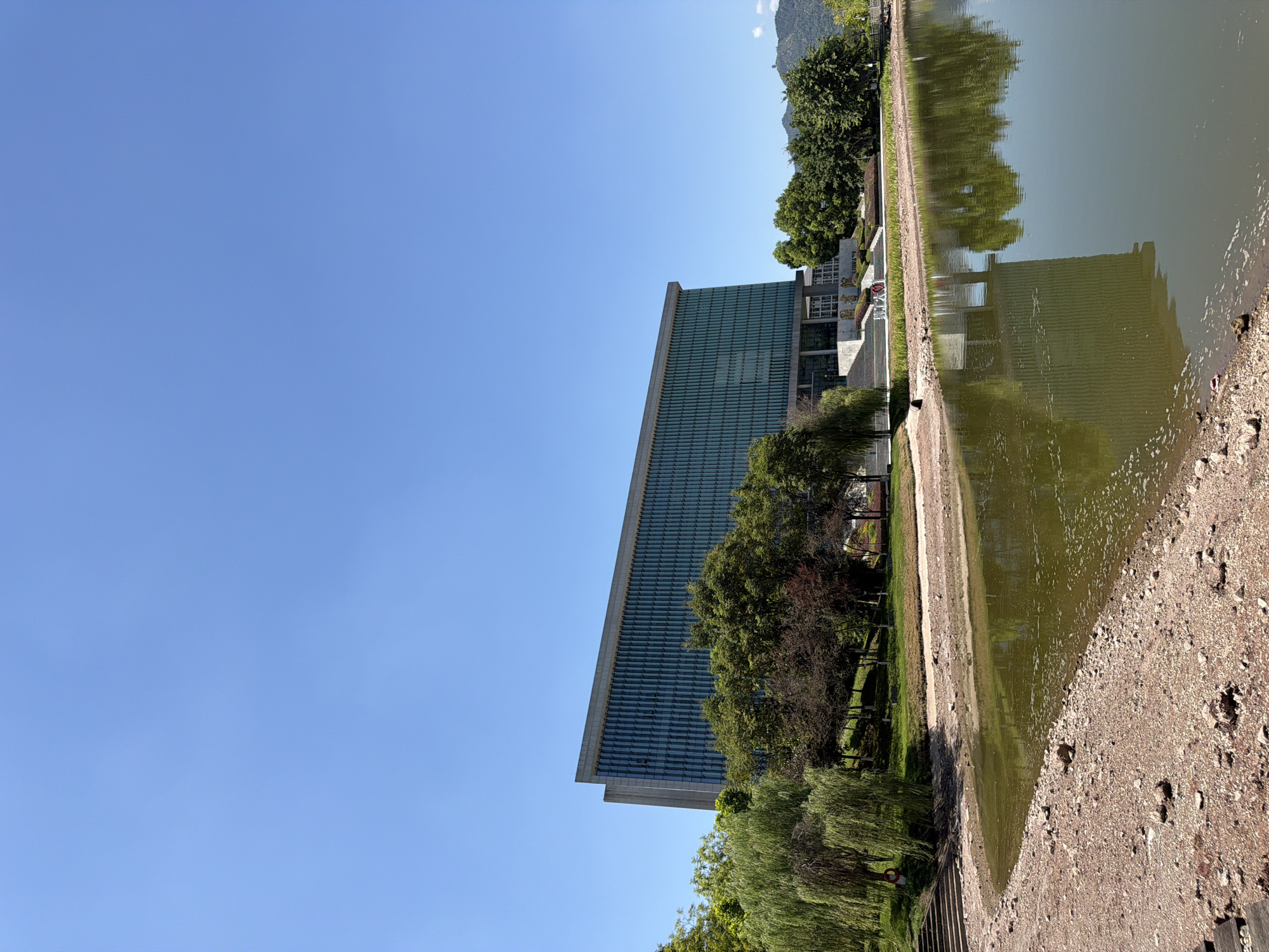 This is a medium-shot, low-angle, eye-level, outdoor, daylight, candid photograph of a modern glass-facade building with a flat roof, likely an educational institution or office building, situated in the city of Jiangjin, China. The building is centrally positioned in the frame and features a long, horizontal structure with numerous vertical glass panels that reflect the clear blue sky. In front of the building, a body of water, possibly a lake or pond, occupies the lower half of the image. The water's surface is slightly rippled and reflects the building and surrounding trees. The shoreline on the left is composed of gravel and small rocks, with a pathway and some steps leading up to a grassy embankment. Lush green trees, including weeping willows and other deciduous varieties, are scattered along the embankment and around the building. The sky is a vibrant, cloudless blue. No people are visible in the image, and there are no explicit signs or landmarks indicating the specific location within Jiangjin, China, beyond what can be inferred from the architectural style and landscaping.