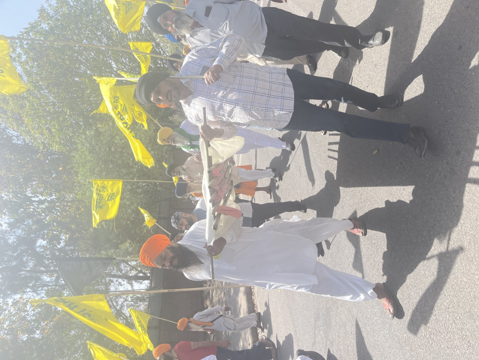 A group of men, all wearing turbans, walk down a paved street in Sahibzada Ajit Singh Nagar, India. They are carrying a large, white cloth stretcher with a wooden frame. The stretcher is held up by two men, with a third man walking behind them. The other men in the procession are carrying yellow flags. The men appear to be in a somber mood, perhaps attending a funeral or a religious procession. There are trees and buildings in the background, and the street is surrounded by a low brick wall. The ground is mostly paved with concrete or asphalt, and there is a slight haze in the air. The scene takes place on a sunny day.