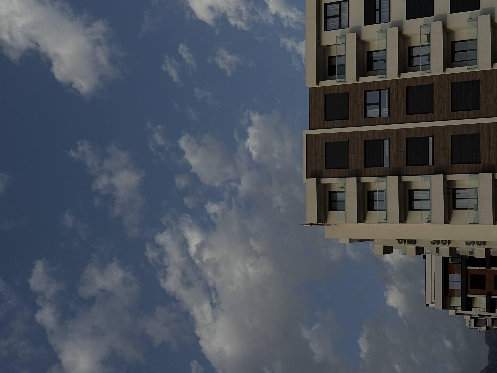 The image presents an upward-angled view of a multi-story building against a backdrop of a partly cloudy sky. The building is characterized by a modern architectural design, featuring a light-colored facade punctuated by dark, rectangular window openings. Some windows have clear glass balcony partitions. The building appears to have horizontal divisions with differing facade treatments. Below the primary building mass is a smaller structure with similar window arrangements, suggesting the building is part of a larger architectural complex. The sky is visible in the background and on the left, with scattered clouds.