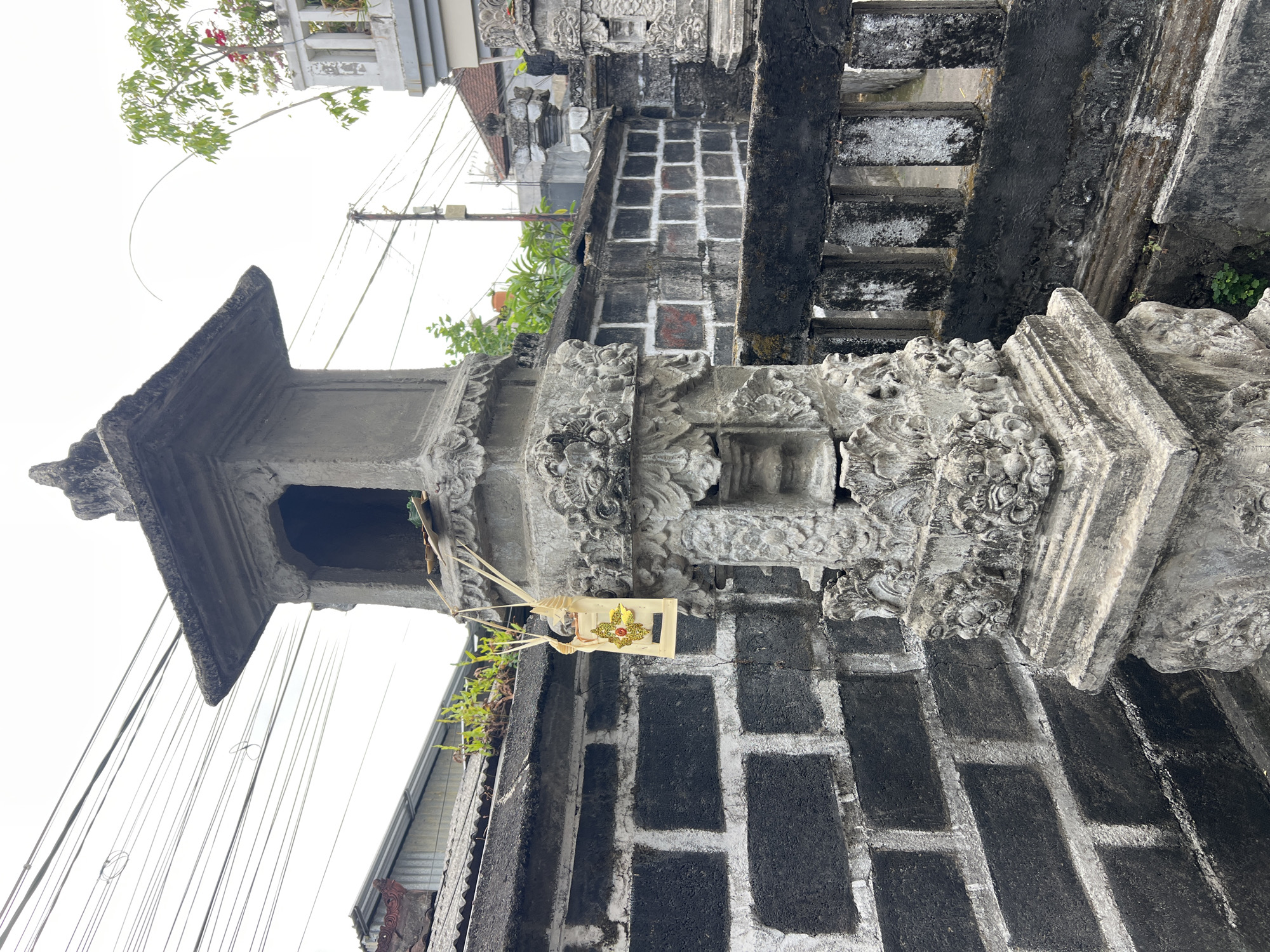 The image shows a close-up of a stone carving on a wall, possibly in Denpasar, Indonesia. The carving is ornate and appears to be part of an outdoor shrine or temple structure. A small, decorative offering is hanging from the structure. In the background, a dark brick wall is visible, along with some electrical wires and a tree. The sky is overcast. The scene appears to be undisturbed, with no people or animals present. The overall impression is one of a religious or cultural site, possibly a place of worship or a traditional Balinese home.