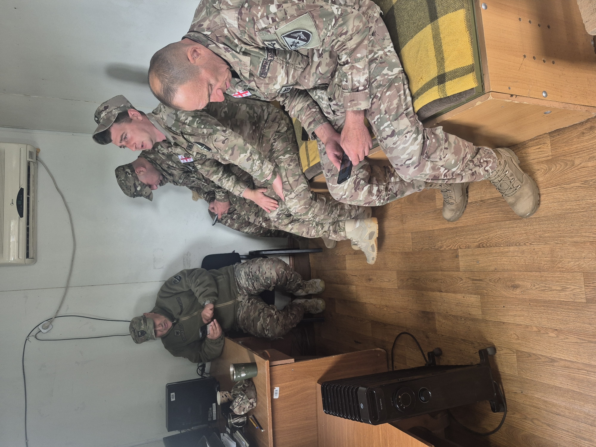Inside a sparsely furnished room with wood-patterned flooring and off-white walls, four soldiers are present. Three soldiers are arranged in a row, leaning against a wooden structure covered with a yellow and green plaid blanket. All three are wearing camouflage uniforms with Georgian flags and patches. The first soldier is looking at a mobile phone he holds in his hands. The remaining two appear to be watching. The fourth soldier sits at a wooden desk, also wearing a camouflage uniform and cap. The desk features a computer monitor, a tin can, a camouflage cap, a coffee cup, and writing utensils. To the right of the desk is a black electric heater. An air conditioning unit is mounted on the wall above and to the left of the soldiers. The location is Tbilisi, Georgia, as evidenced by the Georgian flags on the soldiers' uniforms.
