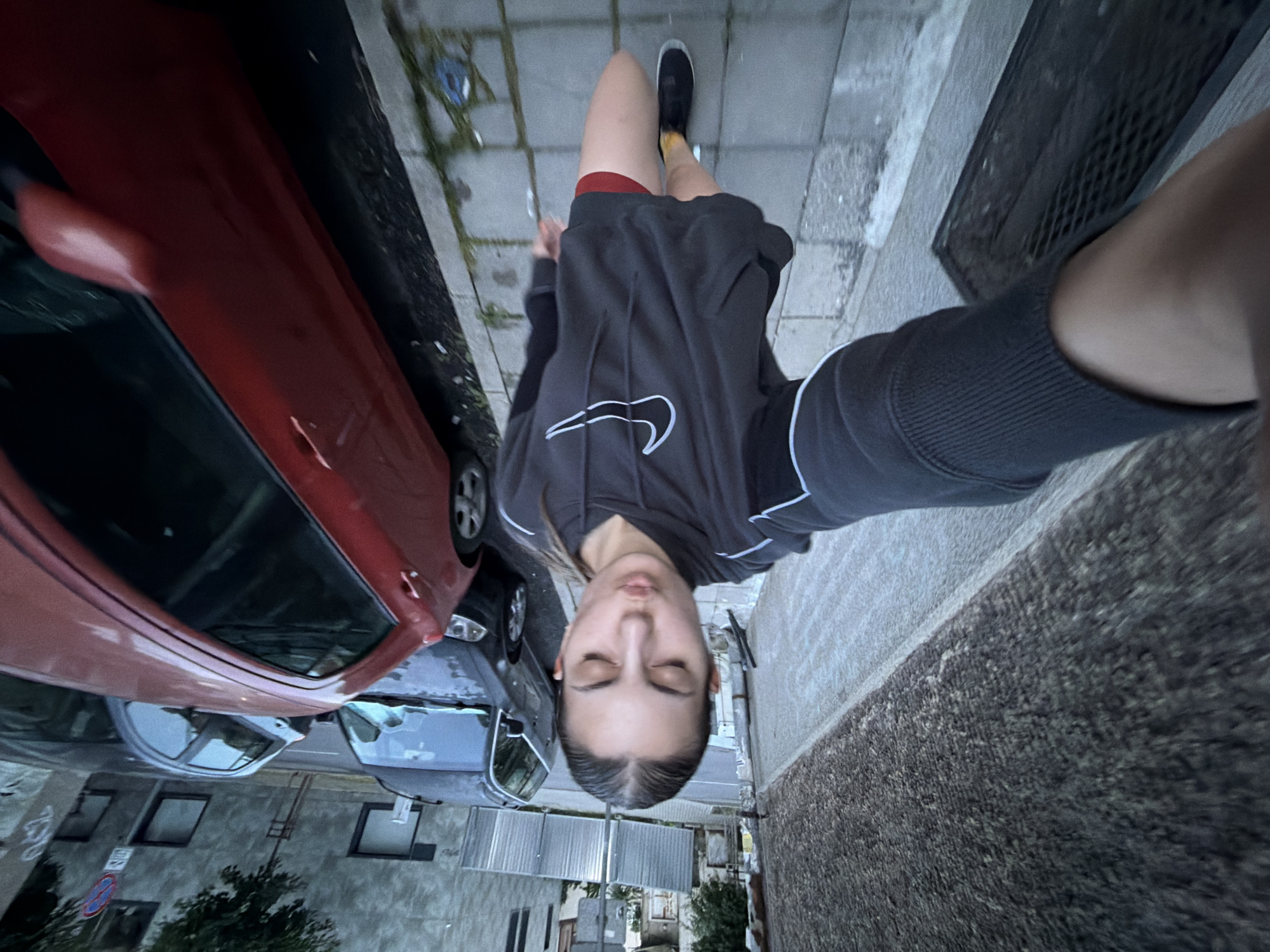 The image captures a unique, overhead selfie taken from a high angle, featuring a person standing on a sidewalk in an urban setting identified as Burgas, Bulgaria. The camera is positioned directly above the subject, resulting in a wide-angle, slightly distorted perspective where the person's head appears more prominent than the body.
The primary subject is a young person, likely female, with dark hair pulled back, seen looking downwards with eyes closed. She is wearing a dark gray hooded sweatshirt adorned with a distinctive white Nike swoosh logo on the chest. Beneath the sweatshirt, red shorts are visible, paired with dark sneakers and bright yellow socks. Her right arm is extended, holding the camera, while her left arm is visible along the right edge of the frame.
The surrounding scene depicts a typical street environment. To the left, a vivid red car is prominently parked along the curb, partially obscuring other parked vehicles further down the street. The subject stands on a gray paved sidewalk, which features some subtle discolorations and possibly moss. To the right, a dark metal grate, likely a storm drain cover, is embedded in the pavement. In the background, an assortment of urban buildings with varying architectural styles and windows line the street. A blue and white circular road sign, characteristic of European traffic signs (e.g., "no waiting" or "no stopping"), is partially visible amidst the buildings. Sparse green foliage from trees or bushes adds a touch of nature to the otherwise concrete landscape.
The lighting suggests an overcast day, as there are no harsh shadows, contributing to a soft and diffused illumination of the scene. The overall mood is casual and contemplative, capturing a personal moment within a bustling urban backdrop. The most prominent visible text is the "Nike" logo on the sweatshirt.