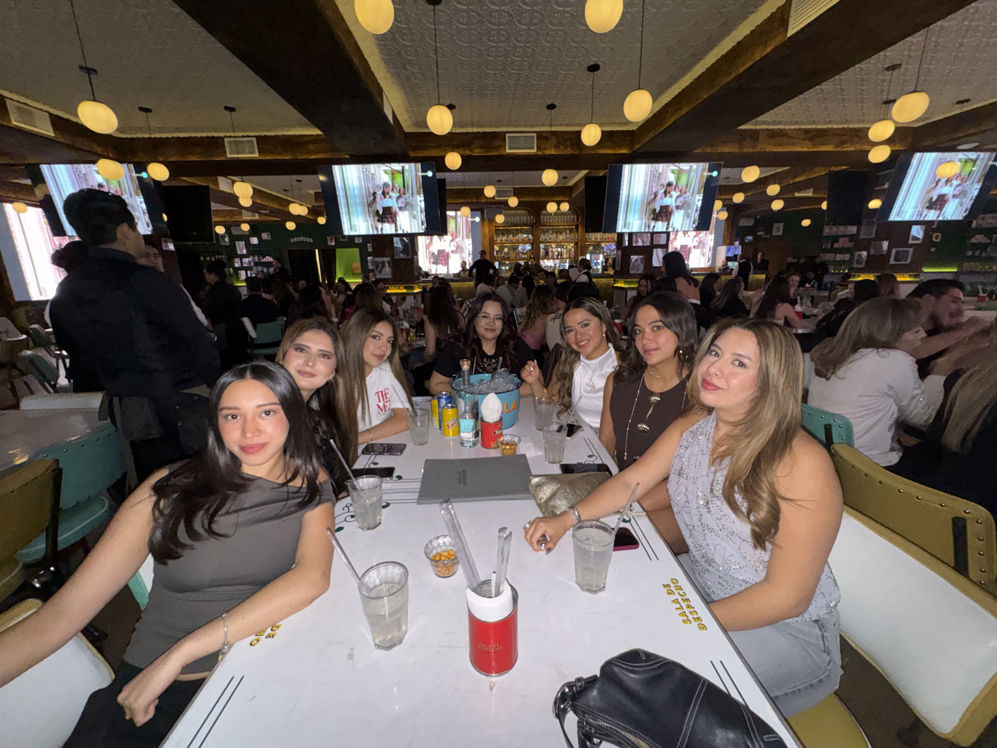 The image portrays an interior scene within Sala de Despacho, a restaurant and bar situated at Av. Rosendo Marquez 1122, La Noria, 72410 Puebla, Pue., Mexico. Seven young adult women are seated around a white rectangular table in the foreground, facing the camera. The woman on the far left wears a dark gray off-the-shoulder top, while the woman on the far right wears a light blue, sequined halter top and gray shorts. Other women at the table wear casual attire including white t-shirts, white blouses, and dark tops. The table is occupied by multiple clear glasses containing beverages and straws, a red cylindrical container holding additional straws, a small bowl of peanuts or similar snacks, a gray menu booklet, a black purse, a gold clutch bag, and several mobile phones.
The restaurant's interior features a dark wooden beamed ceiling with decorative white pressed tin panels and numerous hanging spherical yellow light fixtures. Large flat-screen televisions mounted on the walls display what appears to be a performance with individuals in kilts. A fully stocked bar, complete with bottles and a visible bartender, is present in the background. Other patrons are seated at various tables throughout the establishment, some on green booth seating. A male figure in a black jacket stands on the far left, with his back to the camera. The walls are dark green, adorned with framed pictures and a partially visible neon sign above the bar.