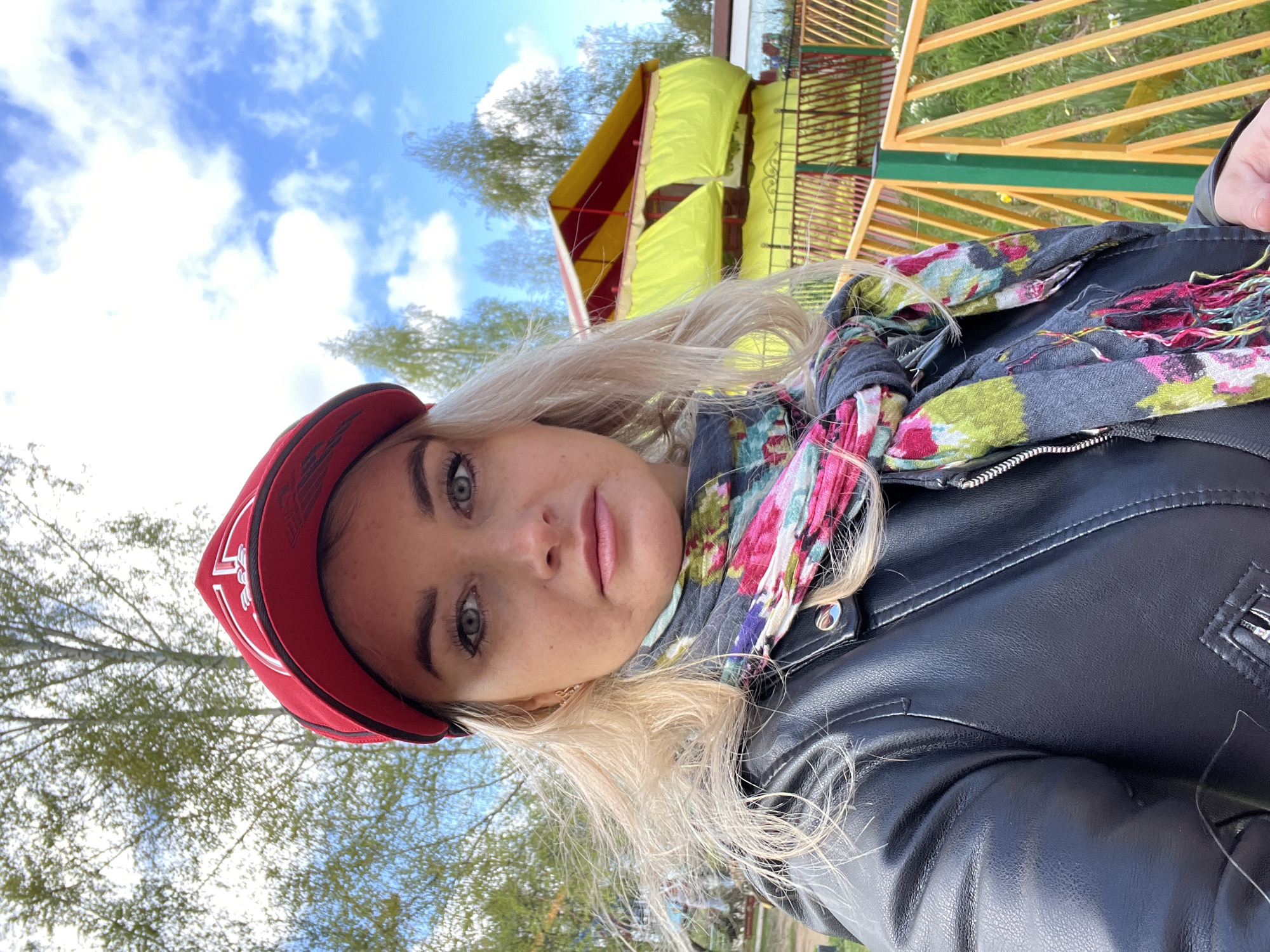 A selfie of a fair-skinned young woman with blonde hair, wearing a red baseball cap and a black leather jacket with a gray floral scarf. She appears to be outdoors. In the blurred background, there are green trees, a blue sky with white clouds, and a yellow and red structure made of wood, suggesting a park or recreational area. While the precise location within Chaikovsky, Russia, cannot be determined from the image, the presence of the park-like structure indicates an area for recreation or leisure.