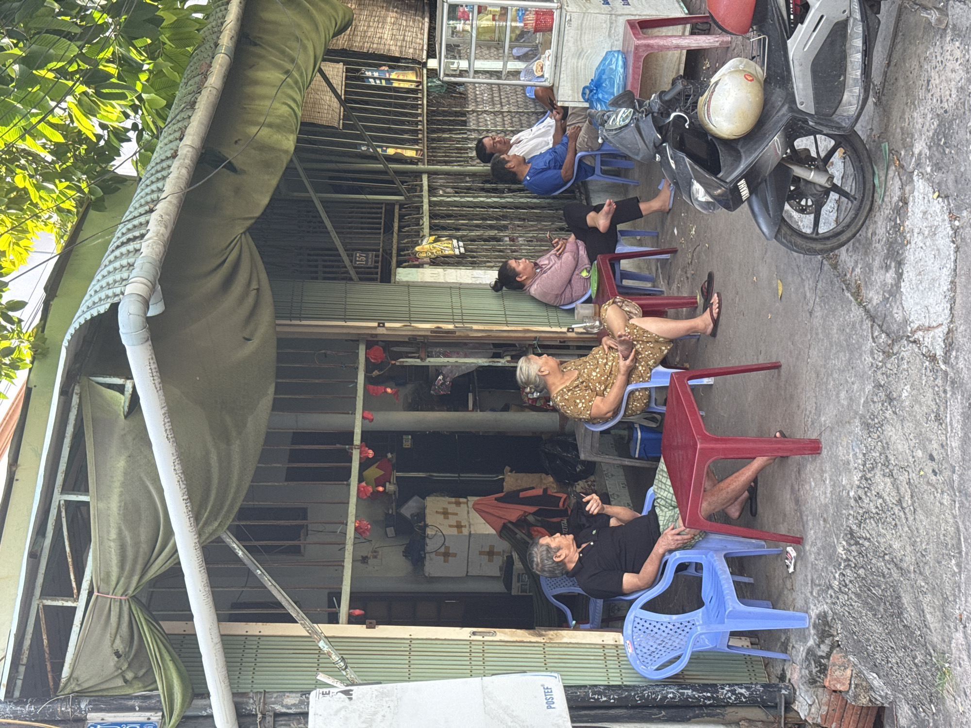 In Phú Thủy, Vietnam, a street scene unfolds in front of a shop with a green, corrugated metal roof and an olive-drab tarp awning. Five individuals are seated on blue plastic chairs around red plastic tables, creating a casual gathering. A man in a black polo shirt and checkered shorts holds a smartphone, while an older woman in a floral dress sits beside him. Further down, a woman with her hair in a bun, wearing a pink shirt and black pants, also looks at her phone. Two men, one in a blue shirt and the other in a white shirt, sit next to her. A motorcycle is parked in the foreground, with a helmet resting on its seat. The shop's entrance is open, revealing stored items and a refrigerator. Red lanterns are visible hanging inside, suggesting a festive or decorative touch. The environment appears to be an urban street, with a concrete sidewalk and a textured, possibly uneven, ground surface.