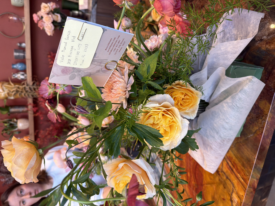 An overhead, slightly angled close-up shot captures a vibrant bouquet of flowers resting on a reddish-brown, marbled surface. The bouquet is primarily composed of large, multi-petaled roses in shades of bright yellow and soft peach, alongside smaller pink and purplish flowers, and various green foliage including some feathery asparagus fern.
A small, rectangular white card with handwritten text is clipped to one of the flower stems using a gold-colored paperclip. The visible handwritten text on the card reads: "Victor Manuel Garcia / 3898 Biscayne Blvd / COSENTINO CITY / Miami, FL 33137". Below this, there is printed text with contact information for a business: "@neroliblume / neroliblume.co / info@neroliblume.com / (786) 967-1887 / 132 NW 27th Street. / Miami 33127". The card itself has a subtle botanical illustration as its background design.
The lower portion of the bouquet is wrapped in light-colored, crinkled tissue paper, with hints of a green floral foam brick visible underneath. In the background, out of focus, more flowers in various shades of pink and red are visible on shelves or in vases, suggesting an indoor setting like a flower shop or market. A person's face, possibly a woman, is partially visible and out of focus in the lower left corner of the image, looking upwards towards the bouquet. The overall lighting suggests an indoor setting with natural light, possibly from a window or overhead.