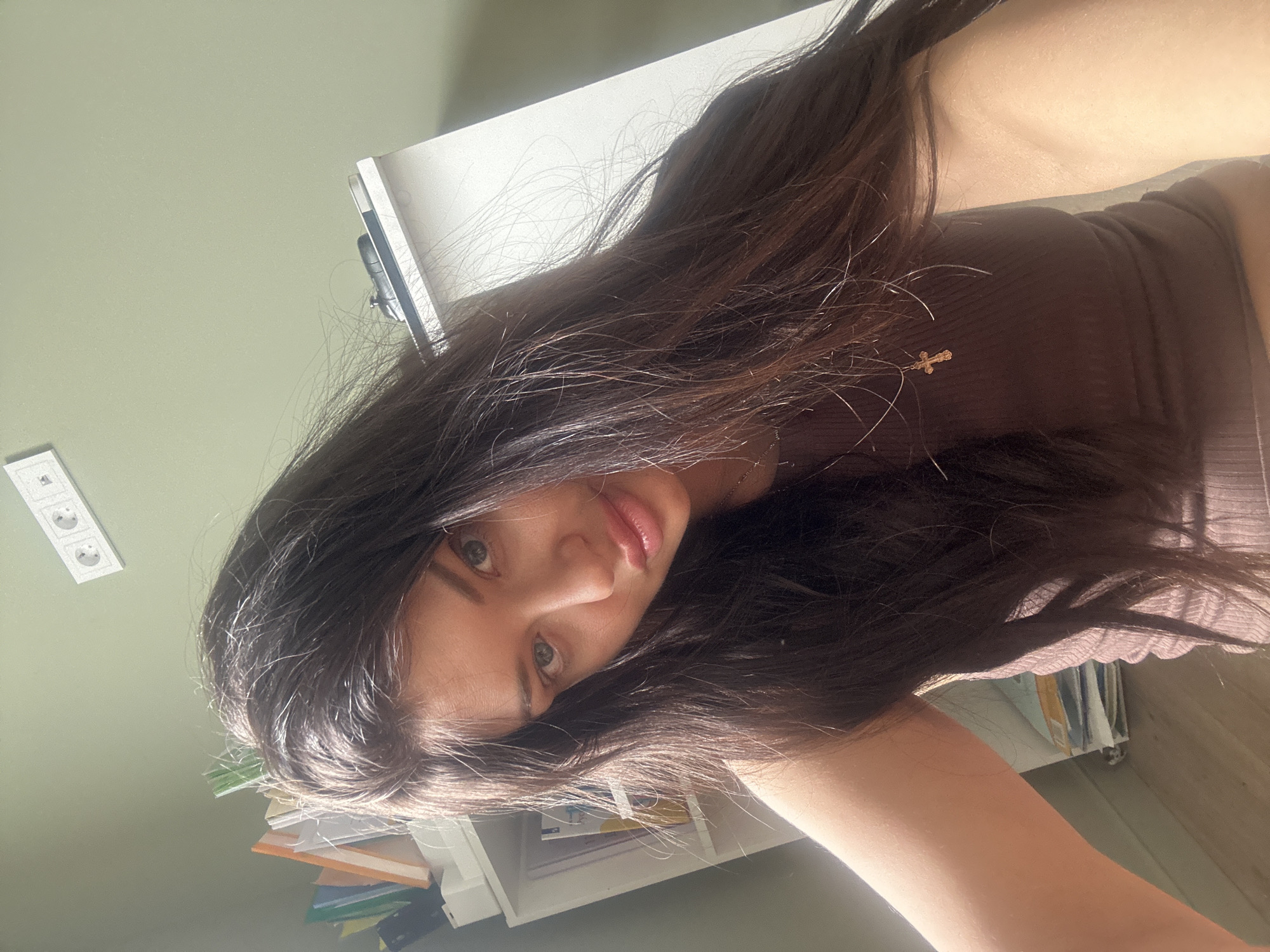A close-up portrait shows a young woman with long, dark brown hair, captured from an overhead or unusual perspective and rotated 90 degrees clockwise. She looks directly at the camera with a neutral expression and slightly parted lips. She wears a dark brown or black ribbed top, and a small, golden cross pendant is visible on her chest. Her fair skin is illuminated by what appears to be natural light, creating highlights on her hair and shoulders.
The background features a light green wall. To the left, a white electrical outlet with three sockets is mounted on the wall. To the right, a white surface, possibly a desk or shelf, is visible, with stacks of books, papers, or folders. The overall setting is indoors, likely a room or office, illuminated by daylight. The scene suggests a casual, personal moment, possibly a selfie, taken in Khimki, Russia. No visible text is present.