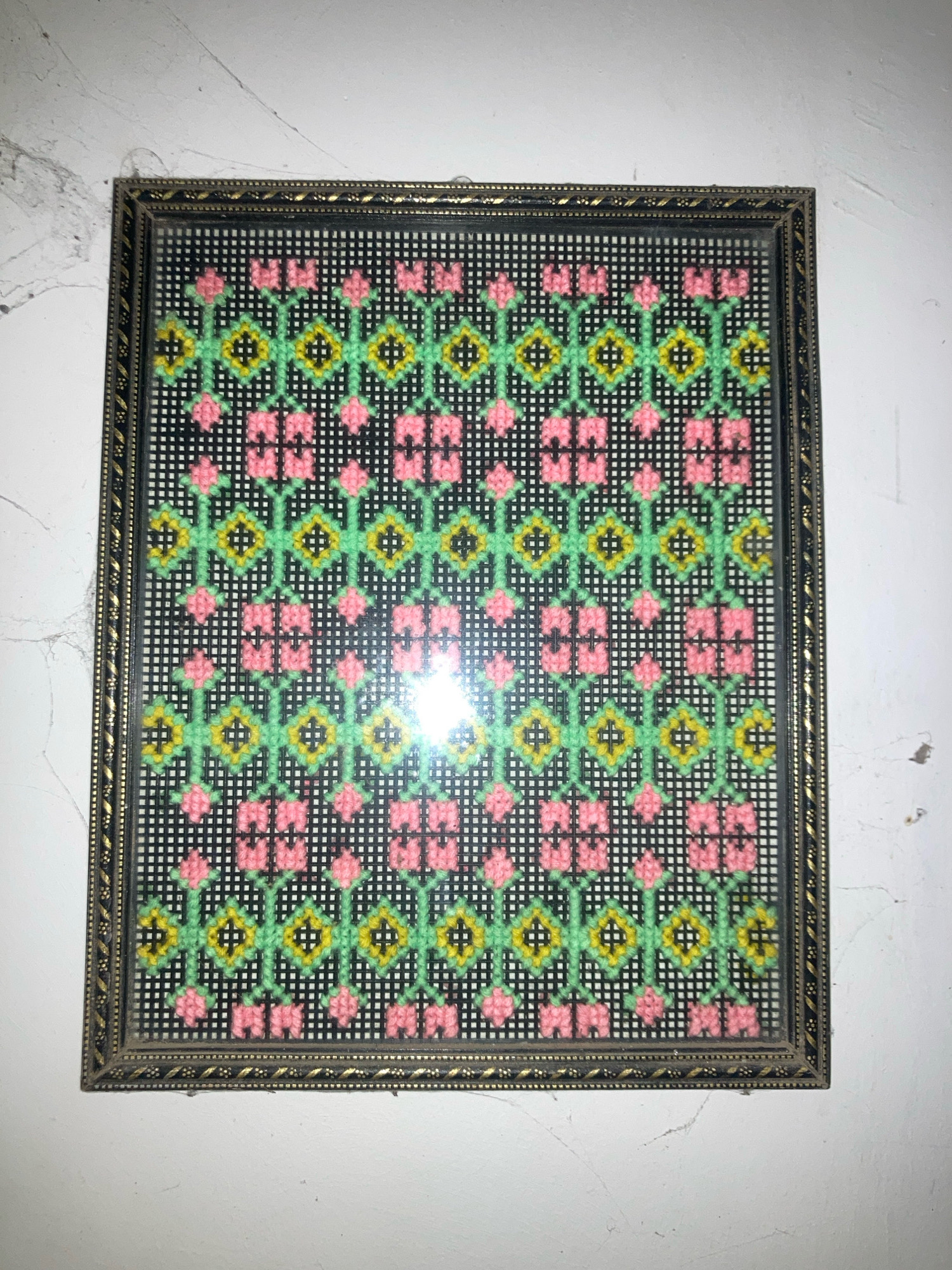 A framed needlepoint tapestry hangs on a white, textured wall. The tapestry features a repeating geometric pattern of pink flowers, green stems, and yellow diamond shapes against a black background. The frame is rectangular with a dark, textured border and a thin, decorative gold trim. The lighting in the image creates a strong reflection on the glass of the frame. The wall appears to be in a domestic setting, possibly a home in Daladagama, Sri Lanka, as indicated by the context.