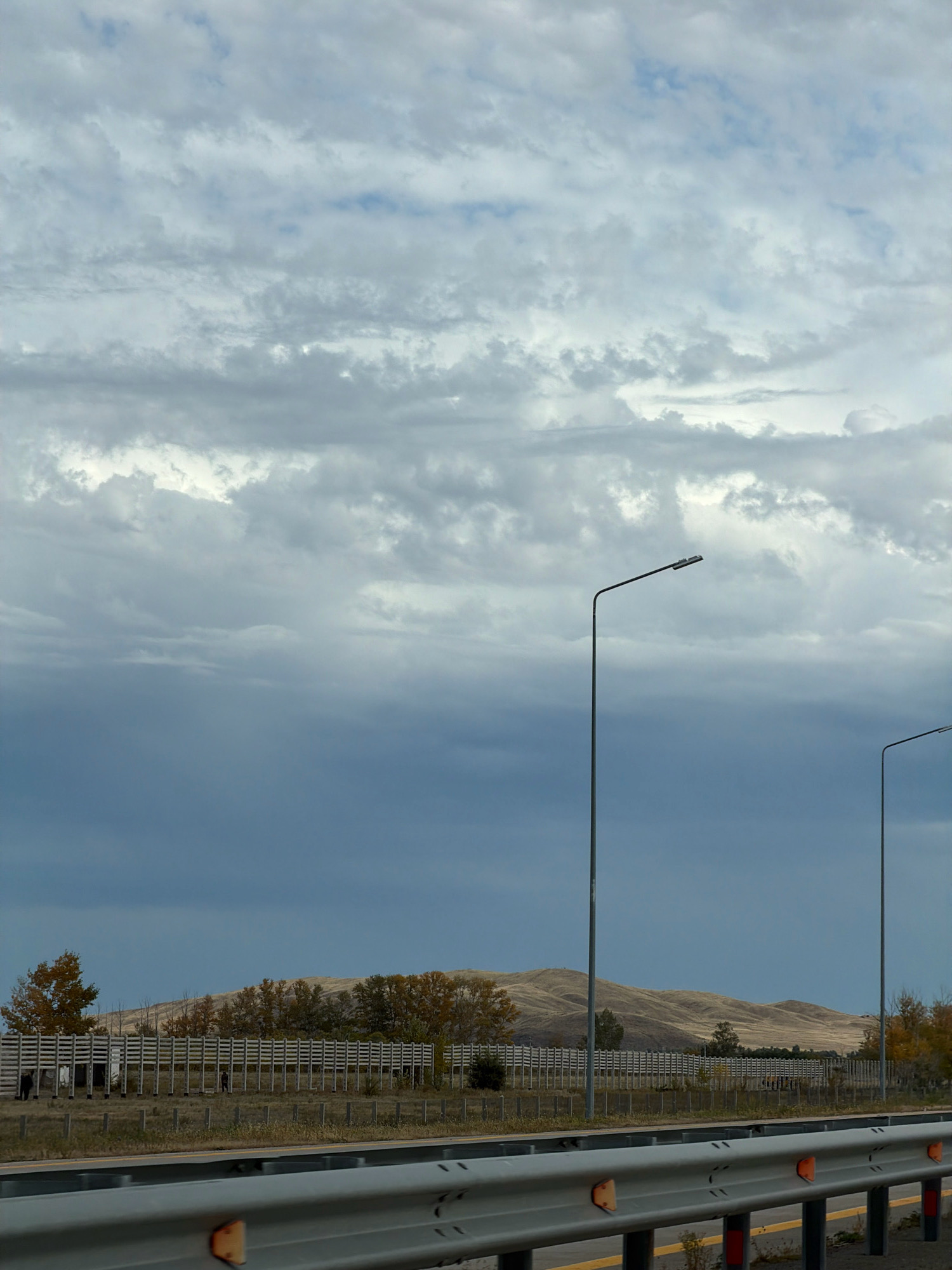 The image depicts an overcast sky with layered clouds over a landscape. In the foreground, a metal guardrail runs horizontally across the lower portion of the frame. Beyond the guardrail, a row of tall, slender lampposts are visible, with two prominently featured. In the middle ground, a long, light-colored wooden fence or barrier stretches across the scene. Behind the fence, a line of trees with autumnal foliage is present, and further back, rolling hills with sparse vegetation form the background. The overall impression is of a somewhat desolate, open area, possibly along a highway. The location is identified as Prirechnoye, Kazakhstan. There are no discernible people or specific actions taking place within the frame.
