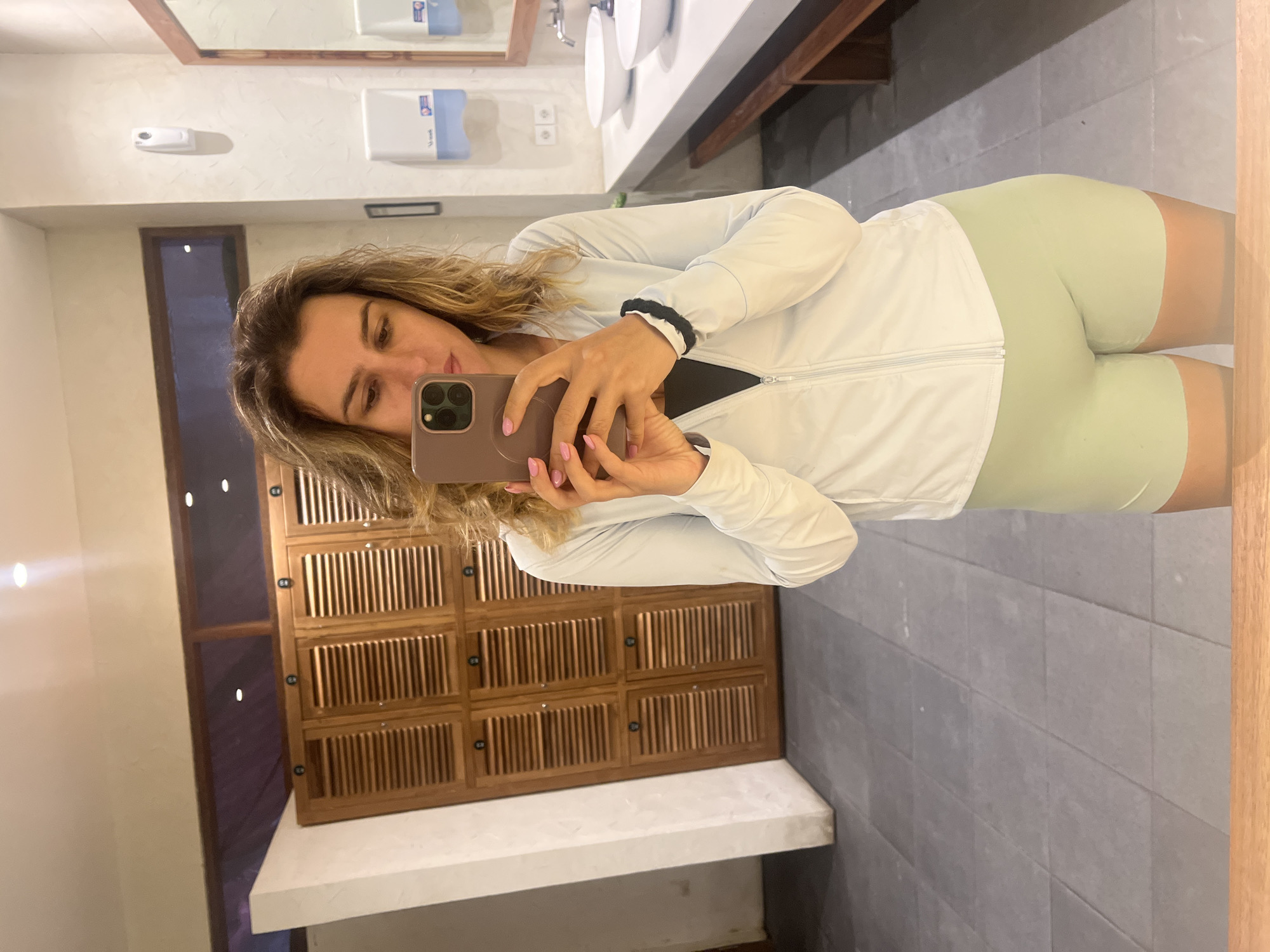A woman takes a mirror selfie in a locker room. She is wearing a light blue zip-up jacket over a black sports bra and light green bike shorts. She has blonde, wavy hair and is holding a pink phone in her right hand, taking the photo. In the background, there are wooden lockers with slatted doors, a tiled floor, and a vanity area with sinks and dispensers. The location is identified as North Kuta, Indonesia.