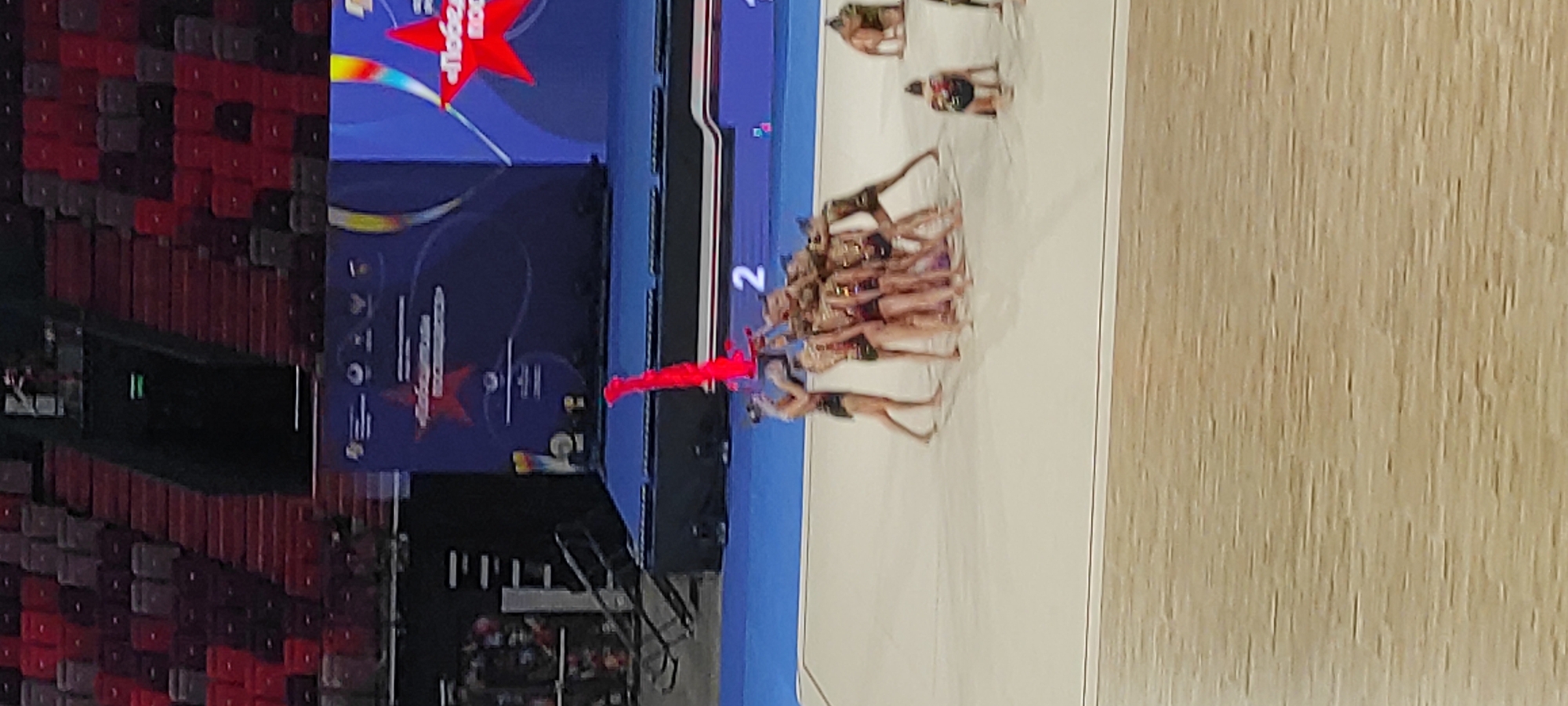 In an indoor arena with tiered red and grey seating visible in the background, a group of young female gymnasts in dark leotards with gold accents are performing on a light-colored mat. The gymnasts are in a complex formation, with several of them supporting others in a pyramid. One gymnast in the center holds a long, bright red ribbon above her head. To the right of the main group, another gymnast is kneeling, and further to the right, two other gymnasts are also in kneeling or low poses. A large screen in the background displays graphics, including red stars and text in Russian, indicating a competition. A numerical display showing "2" is visible near the gymnasts. The overall scene depicts a moment of high-level performance in rhythmic gymnastics. The presence of the screen with Russian text suggests the event is likely taking place in Russia. Given the prominence of the Cyrillic alphabet and the typical venues for major sporting events of this nature, it is probable this image was captured in Moscow, Russia.