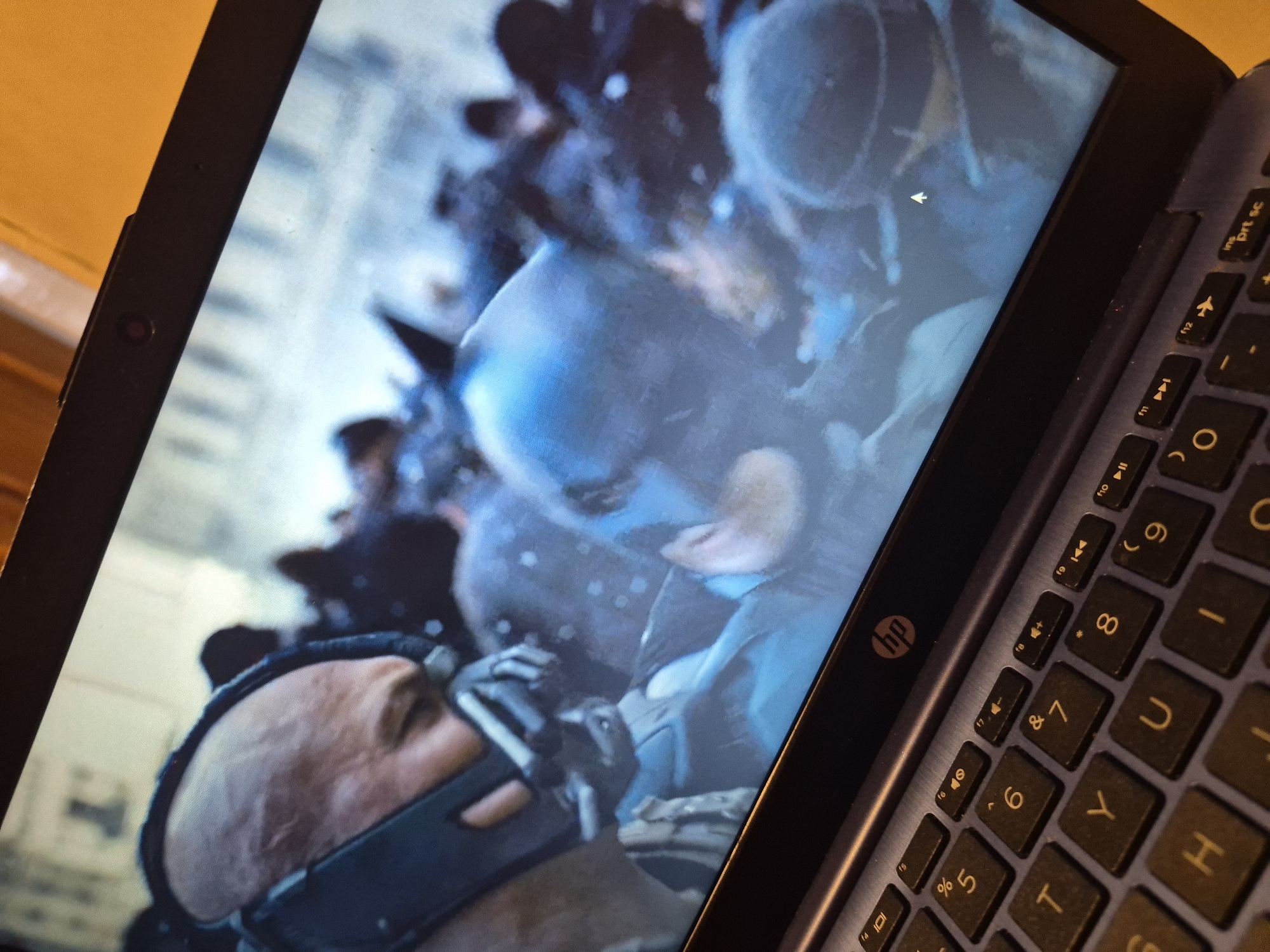 The image shows a laptop screen displaying a scene from a movie, likely Batman: The Dark Knight Rises. In the foreground of the movie scene, a man with a bald head and a distinctive mask covering his lower face, resembling the character Bane, is visible. Behind him, other figures in dark clothing are present, some appearing to wear helmets, suggesting a chaotic or confrontational situation. The background of the movie scene is blurred and indistinct, with hints of urban architecture. The laptop itself is a dark color, with a visible HP logo on the bezel above the screen. The keyboard of the laptop is also visible in the foreground, with a standard QWERTY layout and various function keys. The overall lighting in the image suggests it was taken indoors, with the laptop screen being the primary light source. There is no discernible information within the image that indicates the location in the city of Sandwell, United Kingdom.