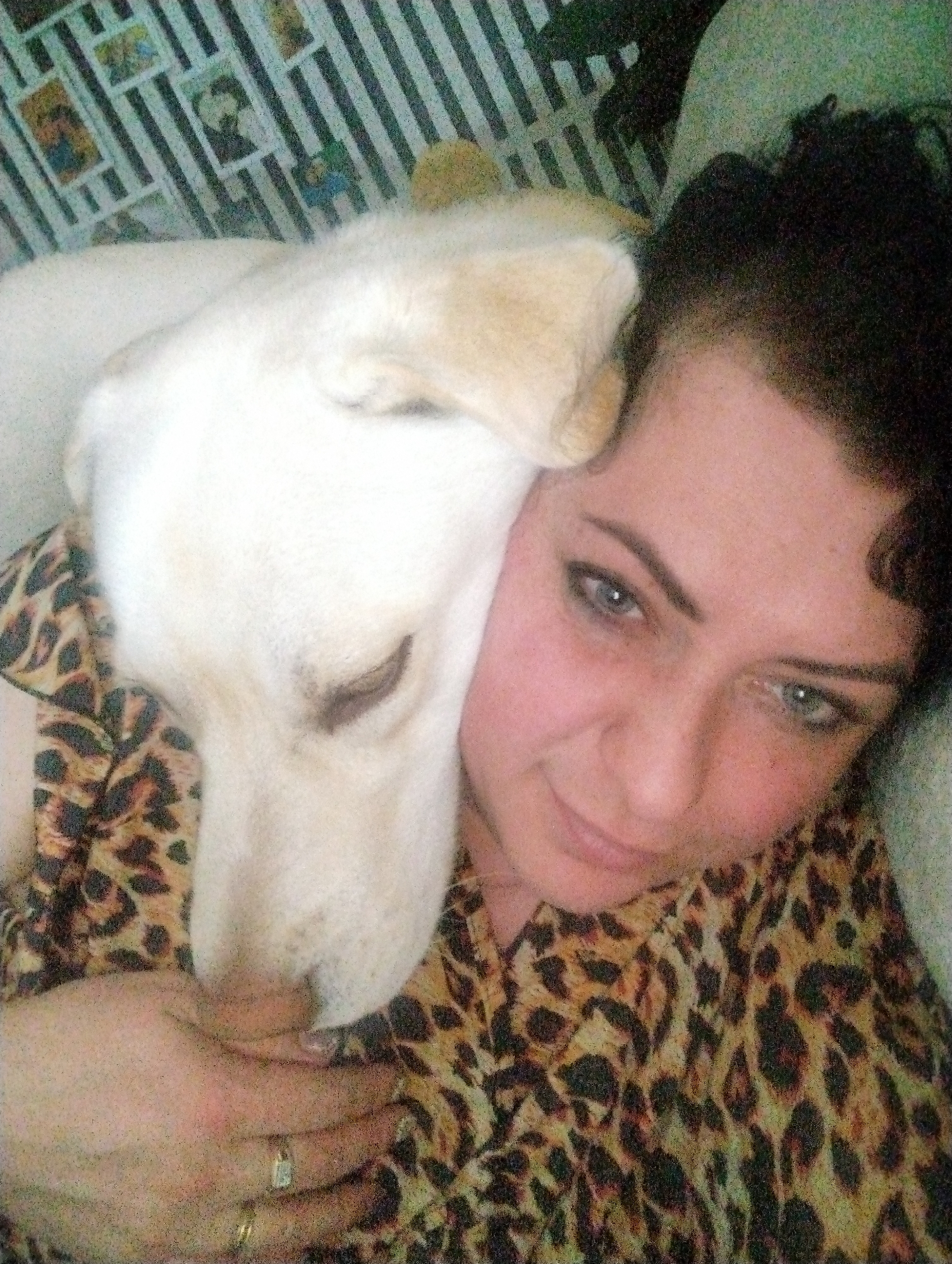 A woman is pictured in a close-up selfie with a white dog. The woman has dark, curly hair, blue eyes, and fair skin. She is wearing a leopard print shirt. The dog is resting its head on her shoulder, with its eyes closed. The background shows a wall with black and white stripes and small images. The location in the city of Grishanki, Russia is not evident from the image.
