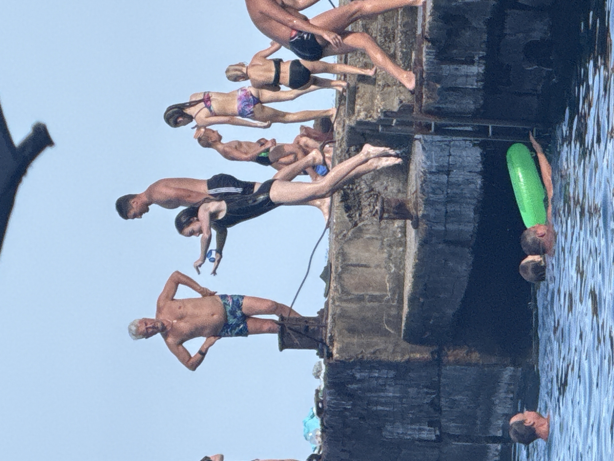 The image captures a scene of people enjoying a day by the water in Partenit, Ukraine. The foreground shows a body of water with several individuals partially submerged. In the middle ground, a group of people are gathered on a concrete structure, likely a pier or breakwater. Some are standing, while others are in motion. A young woman is in mid-air, appearing to be jumping into the water. Another person is holding a green inflatable tube. The background features a clear sky and hints of the surrounding coastal environment. The overall atmosphere is one of recreation and summer activity.