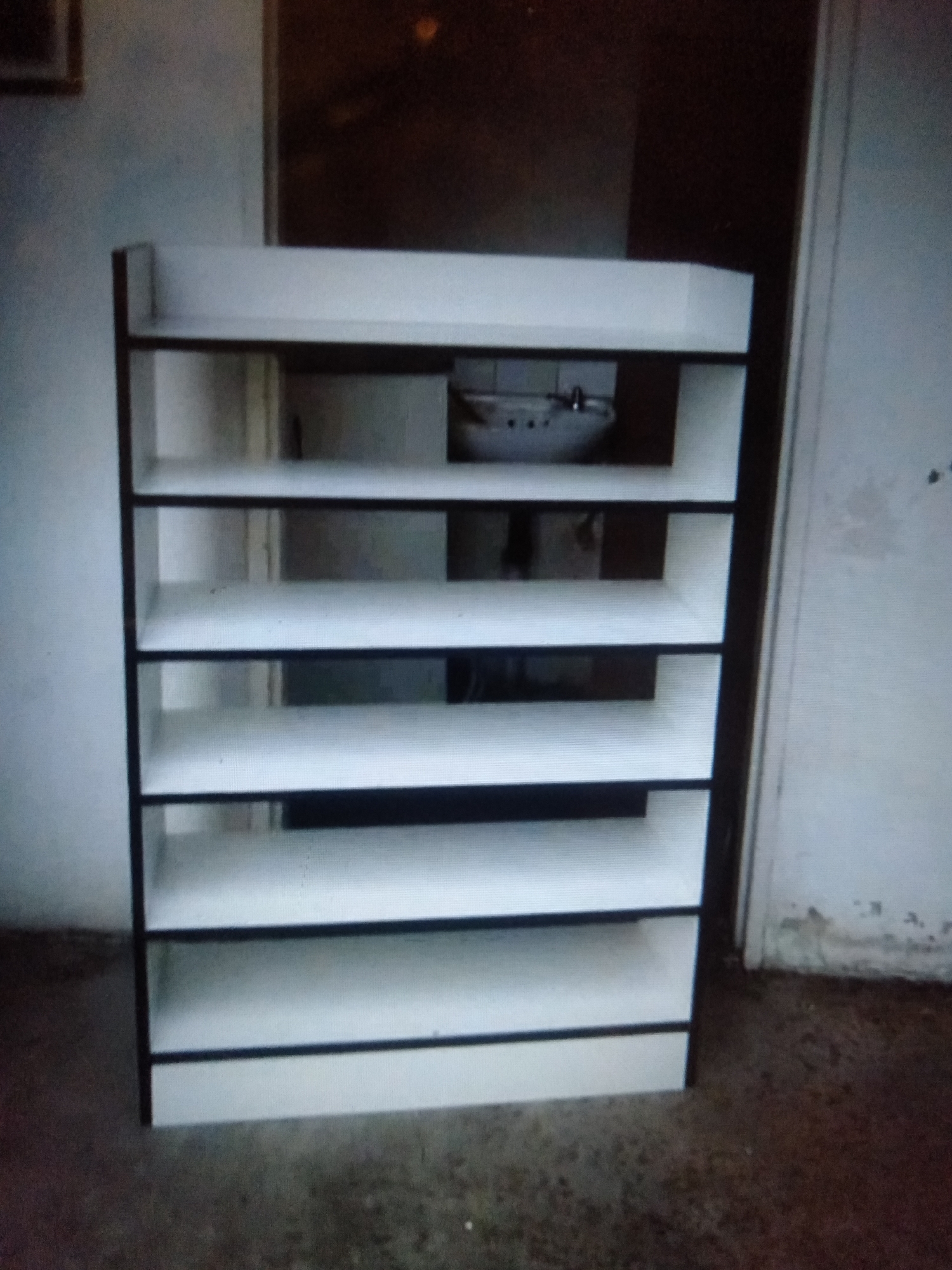 The image shows a white, multi-shelf unit with black trim. The unit appears to be made of wood or a similar material and is standing on a concrete floor. In the background, a sink is visible, suggesting the unit might be in a bathroom or utility area. The lighting in the scene is dim, and the overall atmosphere is somewhat cluttered. The image quality is low, with noticeable pixelation and blurriness, making it difficult to discern finer details. The scene is likely indoors, given the presence of the sink and wall. There is no indication of people or any specific activity taking place. The location context provided is Jalingo, Nigeria, but there are no visual cues within the image to confirm this. No text is visible in the image.