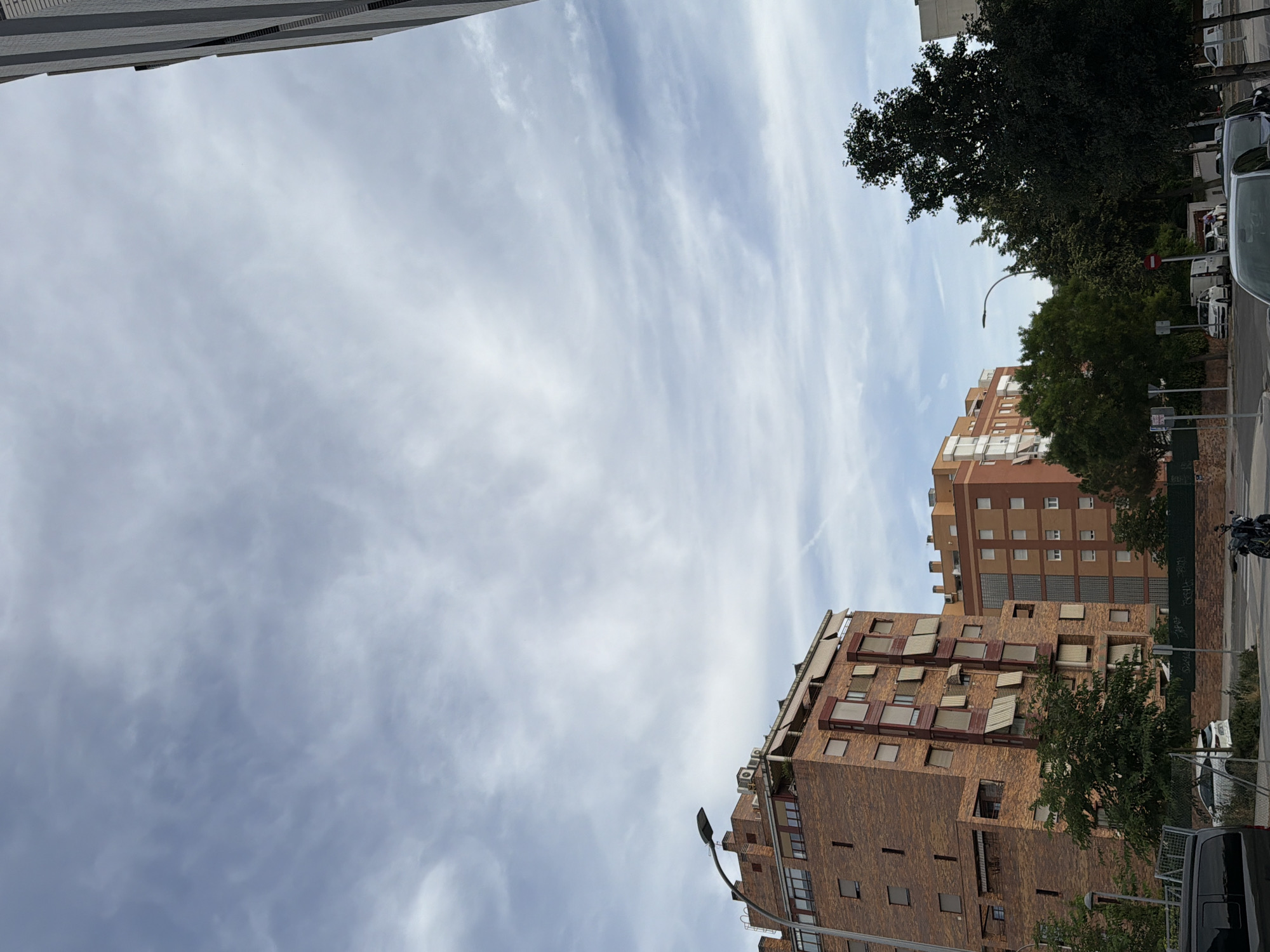 The image captures an upward-looking perspective of a city street. In the foreground, a modern brick building with numerous windows and awnings dominates the lower portion of the frame. To its right, another taller, lighter-colored building is visible. Trees are interspersed between the buildings and along the sidewalk, with some showing full foliage and others appearing to be deciduous.
The sky above is a prominent feature, filled with wispy, white clouds streaking across a pale blue expanse. The sunlight suggests it is daytime, likely mid-morning or afternoon. The angle of the light implies a clear day with some cloud cover.
On the right side of the image, elements of an urban environment are present, including a sidewalk, street lamps, parked cars, and a motorcycle. The architecture of the buildings points towards a residential or mixed-use urban area. There are no visible people in the frame. The overall impression is one of a typical day in a city, with a focus on the interplay between the built environment and the sky.