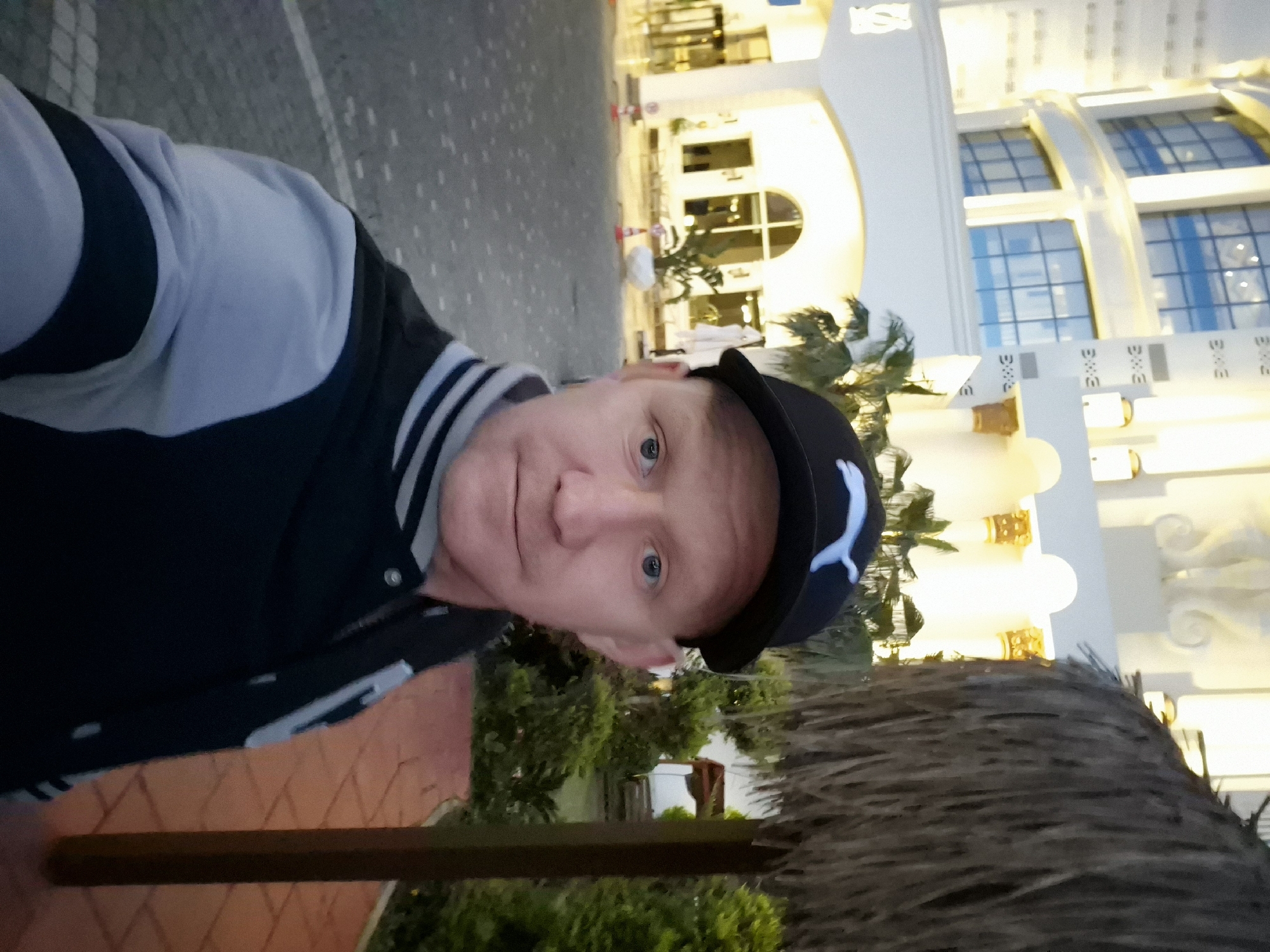 A man wearing a baseball cap and a varsity jacket is taking a selfie. He is standing outside a building with a distinctive arch and ornate details. The building is illuminated, indicating it is dusk or evening. Behind the man is a palm tree and some landscaping. The location is likely Alanya, Turkey, based on the architectural style of the building and the vegetation.