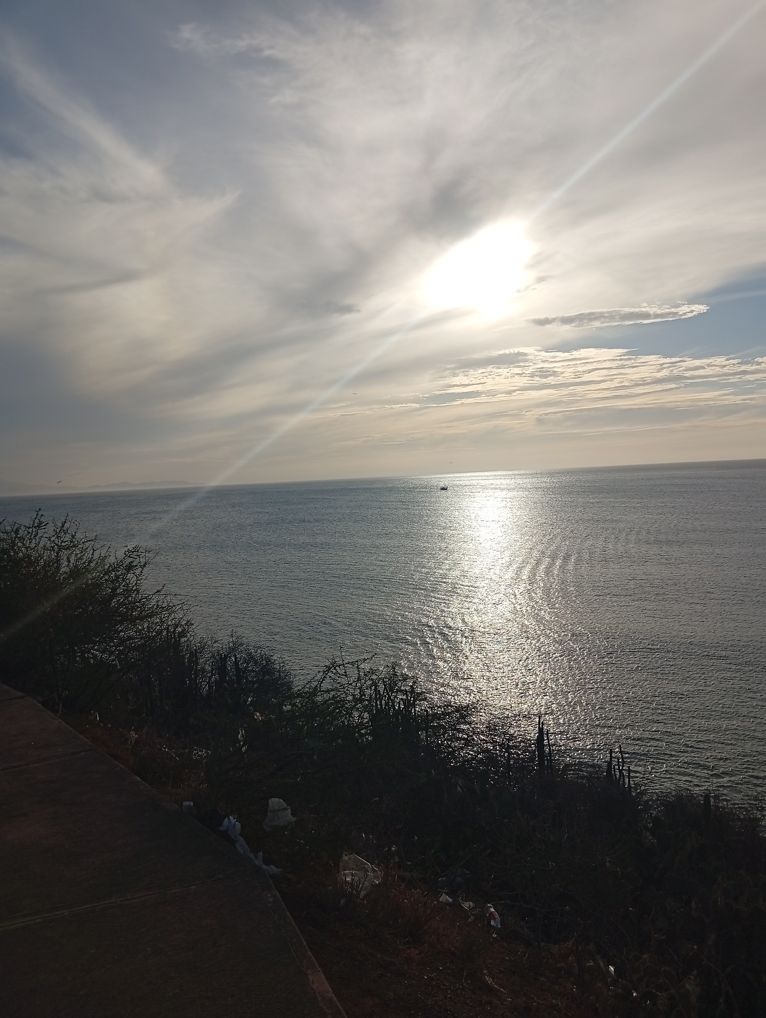 The image shows a seascape at Juan Griego, Venezuela. The scene features a body of water, likely the Caribbean Sea, reflecting sunlight. The sun is obscured by clouds, creating a bright halo effect with visible sun rays. In the foreground, there is vegetation along a roadside or cliffside, with scattered trash visible among the plants. The sky is filled with clouds, with varied textures. A distant landmass is visible on the horizon. There is a single boat in the water.