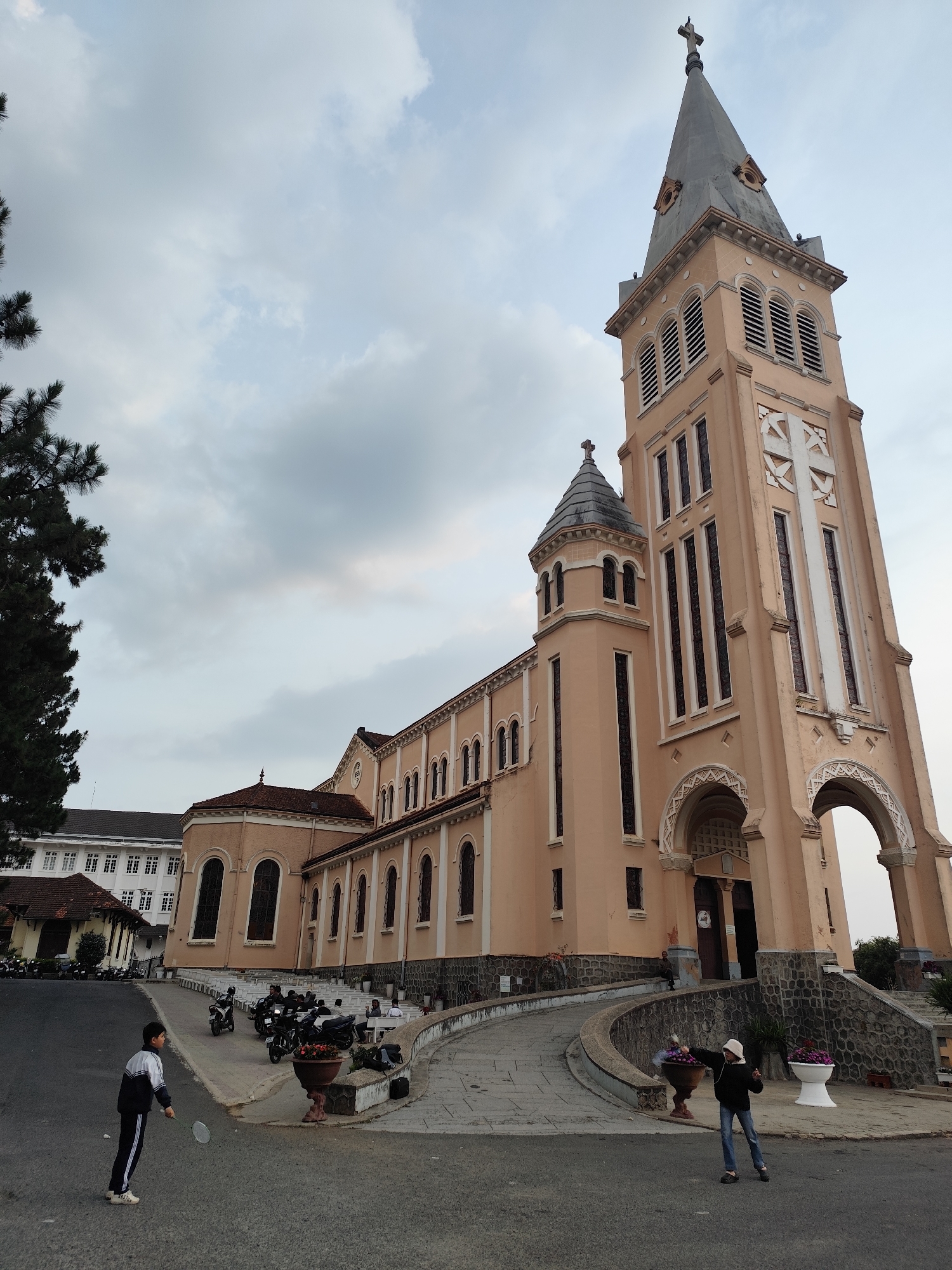 The image depicts the St. Nicholas Cathedral, a Roman Catholic cathedral in Da Lat, Vietnam. The cathedral is a large, pink-beige structure with a tall bell tower topped with a cross. The sky is overcast with gray clouds. In the foreground, a child in a dark tracksuit with white stripes is holding a badminton racket. Across the frame, another person in a dark jacket, jeans, and a white hat is also present. Parked motorcycles are visible on the paved area beside the cathedral. Flowering plants are in pots along the building's walkway. In the background, there is a building with white and brown walls.