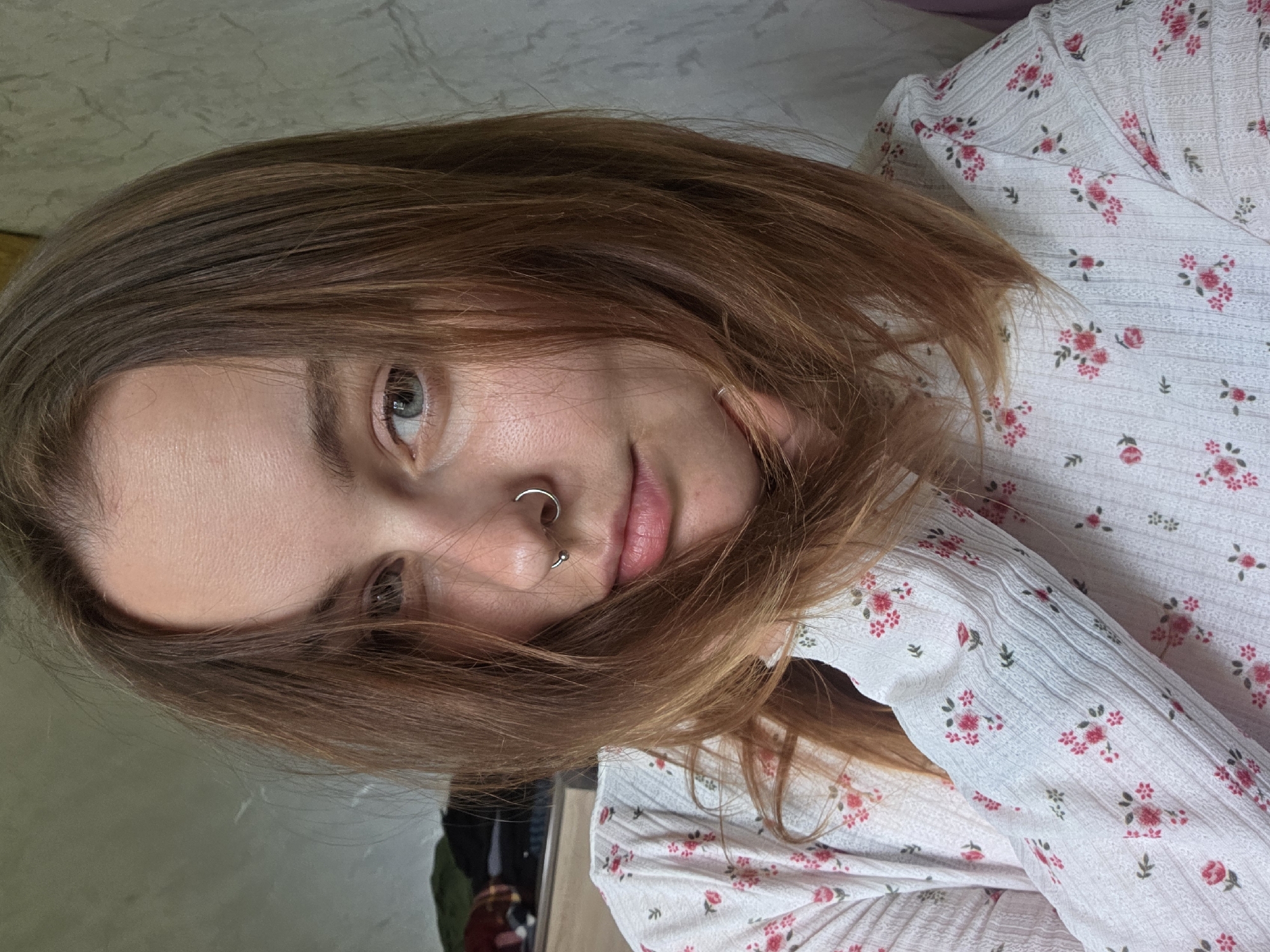 A close-up shot captures a young woman with light brown hair, featuring noticeable auburn highlights. Her skin tone appears fair, and she has a nose piercing. She is wearing a white top adorned with a small pink floral print. The background is blurred and indistinct. The location is unknown.