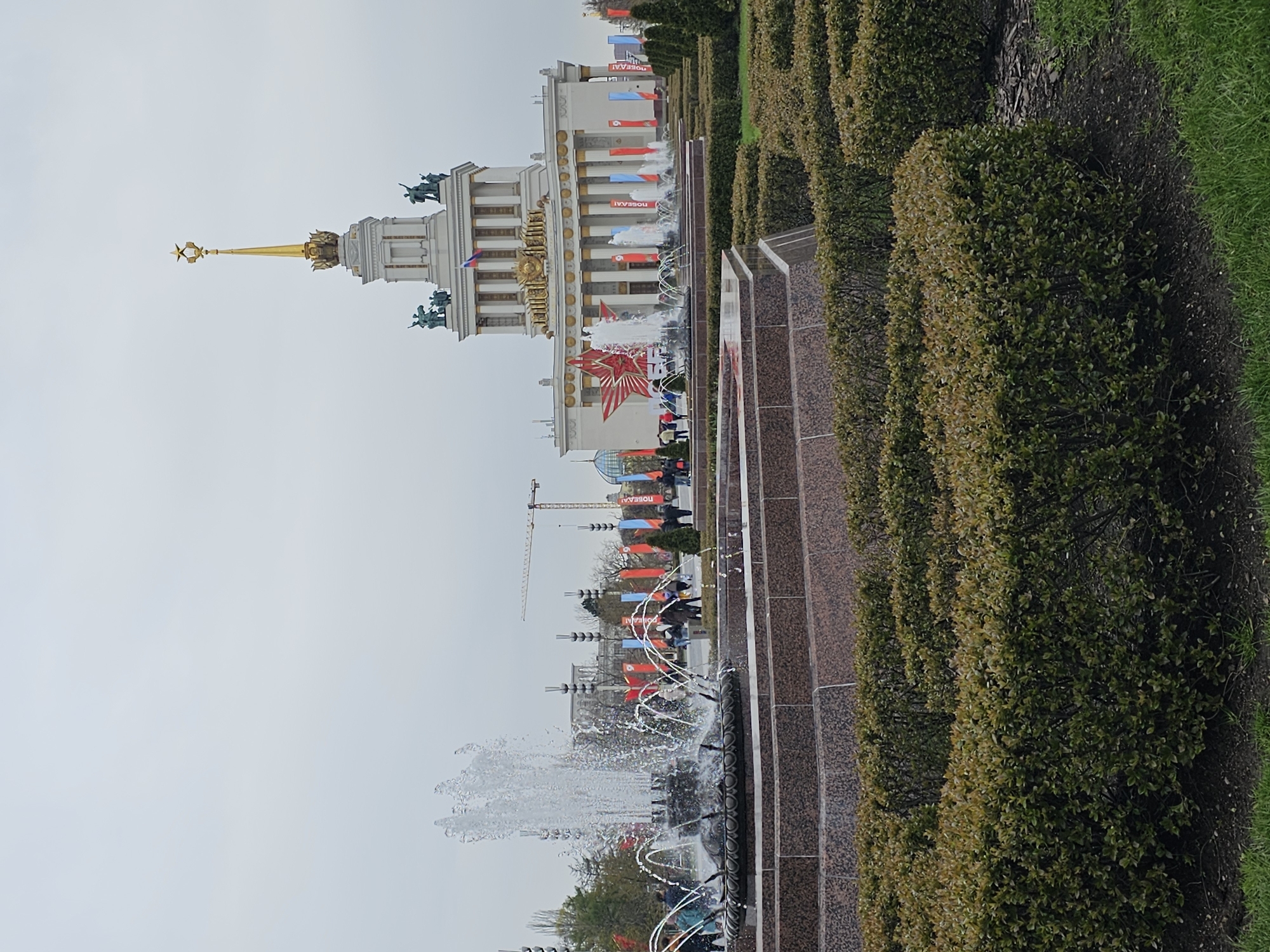 The image depicts the Main Pavilion (Central Pavilion) at the Exhibition of Achievements of National Economy (VDNKh) in Moscow, Russia. The pavilion is a large, ornate structure with a gold spire topped with a star. Banners and flags are displayed on the building. In front of the pavilion is a fountain. Hedges and shrubbery are in the foreground, and a cloudy sky is visible above the pavilion. People are milling around the base of the building. A construction crane is in the background. The image appears to have been rotated 90 degrees counter-clockwise.