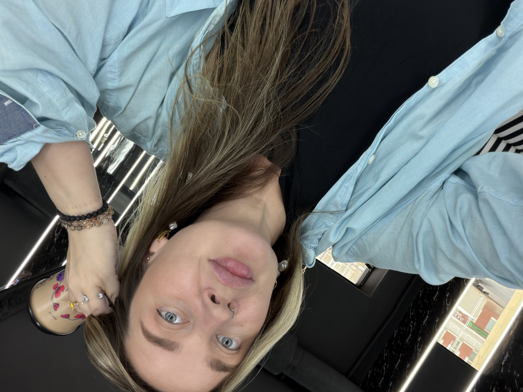 A woman is holding a 400ml tumbler decorated with floral designs. She is wearing a light blue button-down shirt, multiple bracelets on her left wrist, and two rings. Her hair is blonde and brown. She has a nose ring, and her eyes are light blue. She is indoors. Outside the window, there is a building. Based on the Cyrillic text visible on the building across the street, the location is Moscow, Russia.