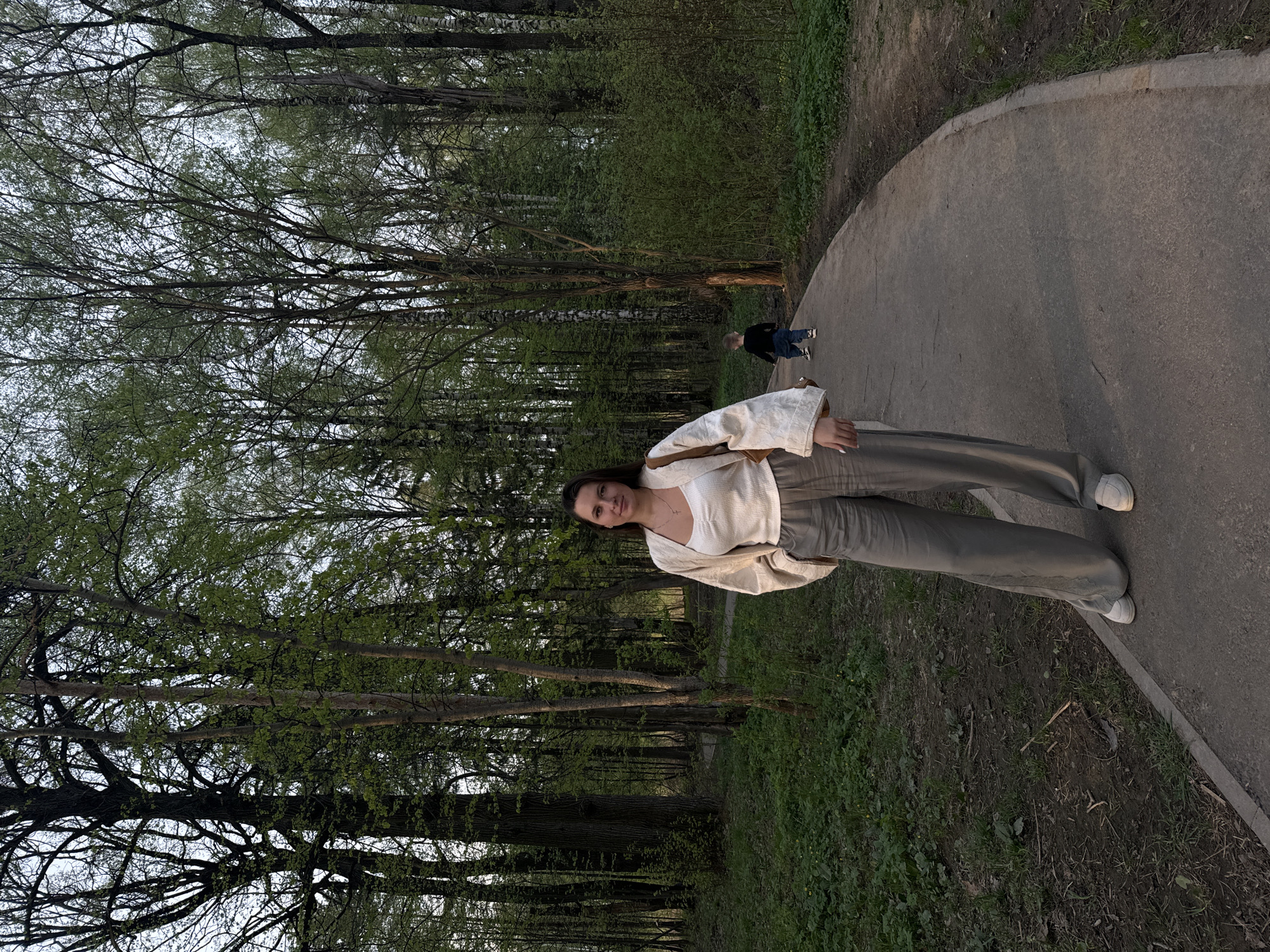 A young woman stands on a paved path in a wooded area, possibly a park. She is wearing light-colored clothing consisting of a cardigan and trousers, and white sneakers. A second person, appearing to be a child, is further down the path in the background. The path curves slightly. Trees surround the path, some with dense foliage, others with bare branches. The location is most likely in the city of Khimki, Russia, based on contextual information.
