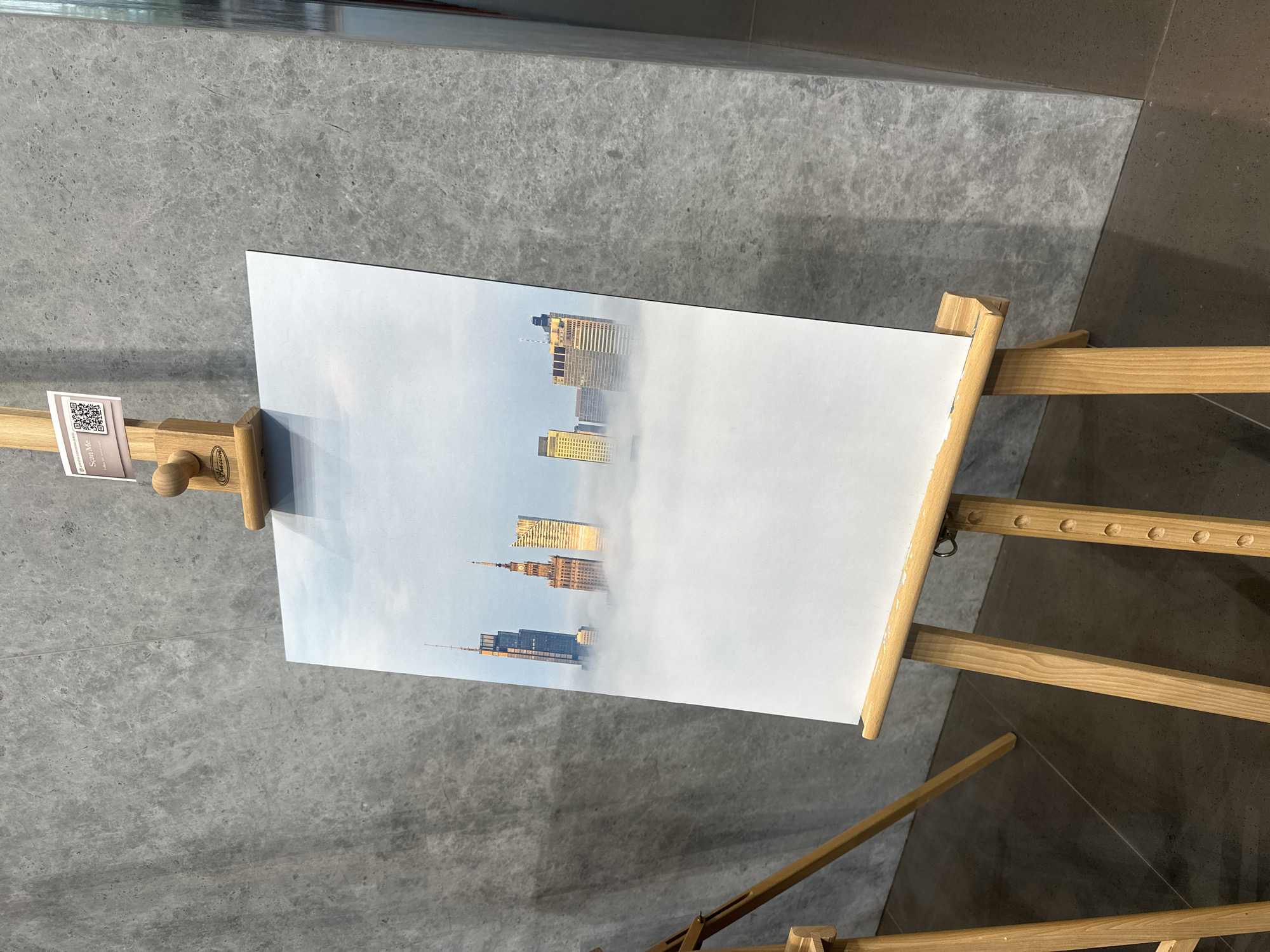 A photograph captures a painting of the Palace of Culture and Science in Warsaw, Poland, displayed on a wooden easel against a textured gray wall. The painting depicts the Palace of Culture and Science emerging from a dense layer of fog, with other skyscrapers visible in the distance. The easel is made of light-colored wood and has an adjustable support for the artwork. A small sign with a QR code and the text "Scan Me" is attached to the top of the easel. The floor is a dark, polished surface, and another wooden easel is partially visible on the left side of the frame.