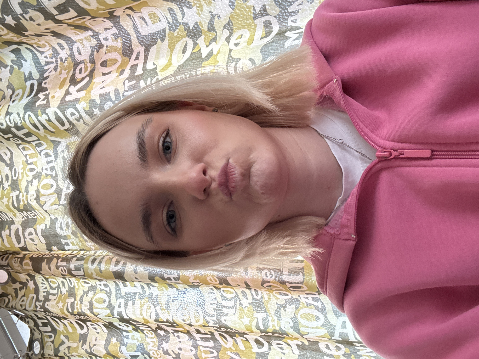 A selfie shot depicts a blonde woman wearing a pink zip-up hoodie and a white t-shirt. She is looking directly at the camera with a slightly puckered mouth. The background consists of patterned curtains with various words printed on them. The location and city are Unknown, Unknown.
