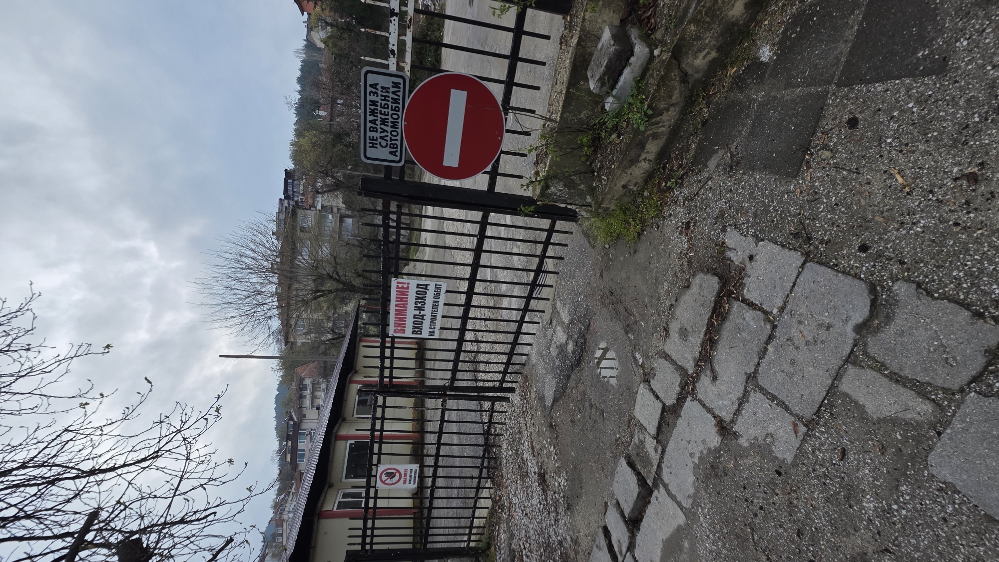 The scene depicts a metal gate at a construction site in Blagoevgrad, Bulgaria. The gate is blocking access to the site and has several signs attached to it. The top sign is a no-entry sign. Below that, there is a warning sign in Bulgarian that reads "ВНИМАНИЕ! ВХОД-ИЗХОД на строителен обект," which translates to "Attention! Entrance-Exit of a construction site." There is also a sign showing a person with a cigarette and a line through it. The ground is paved with concrete blocks. A few trees are near the gate. In the background, buildings and the cloudy sky are visible.