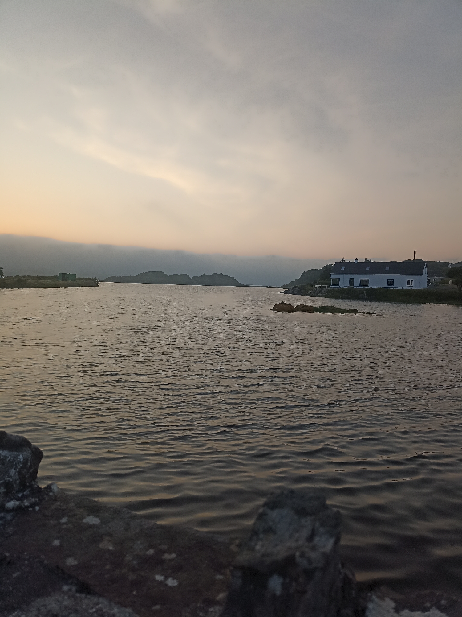 A long shot captures a tranquil body of water, likely a loch or bay, under a pale, overcast sky. The water's surface is gently rippled, reflecting the muted tones of the sky. In the distance, a low-lying, dark green structure, possibly a shed or small building, sits on the left bank. Further to the right, a picturesque white house with a dark roof is nestled on a rise overlooking the water. A small, rocky outcrop emerges from the water in front of the house. The foreground is dominated by the textured surface of a weathered stone barrier, possibly a wall or jetty, with patches of lichen. The overall scene suggests a serene, possibly remote, coastal or lakeside environment in Ireland, captured at either dawn or dusk.