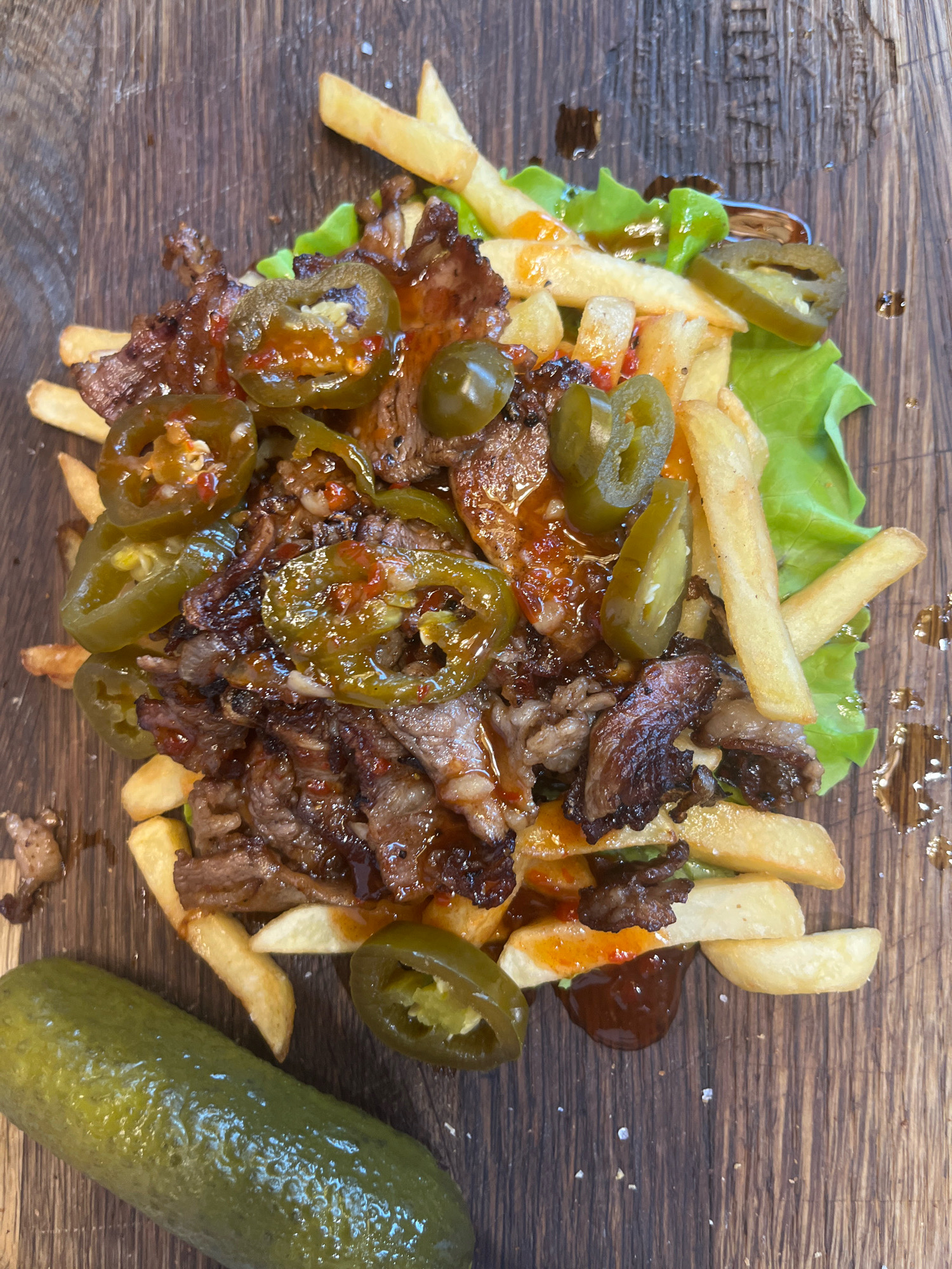 This is a top-down, close-up, eye-level shot of loaded fries with steak and jalapeños served on a wooden surface. The fries are golden brown and appear to be coated in a glossy sauce, interspersed with pieces of thinly sliced steak and bright green jalapeño rings. A large dill pickle lies to the left of the plate, partially out of frame. The wooden surface shows a visible grain pattern and is speckled with what appears to be salt. The lighting is bright, casting minimal shadows. The image does not contain any recognizable landmarks or details that would indicate a specific location within the city of Perm, Russia.