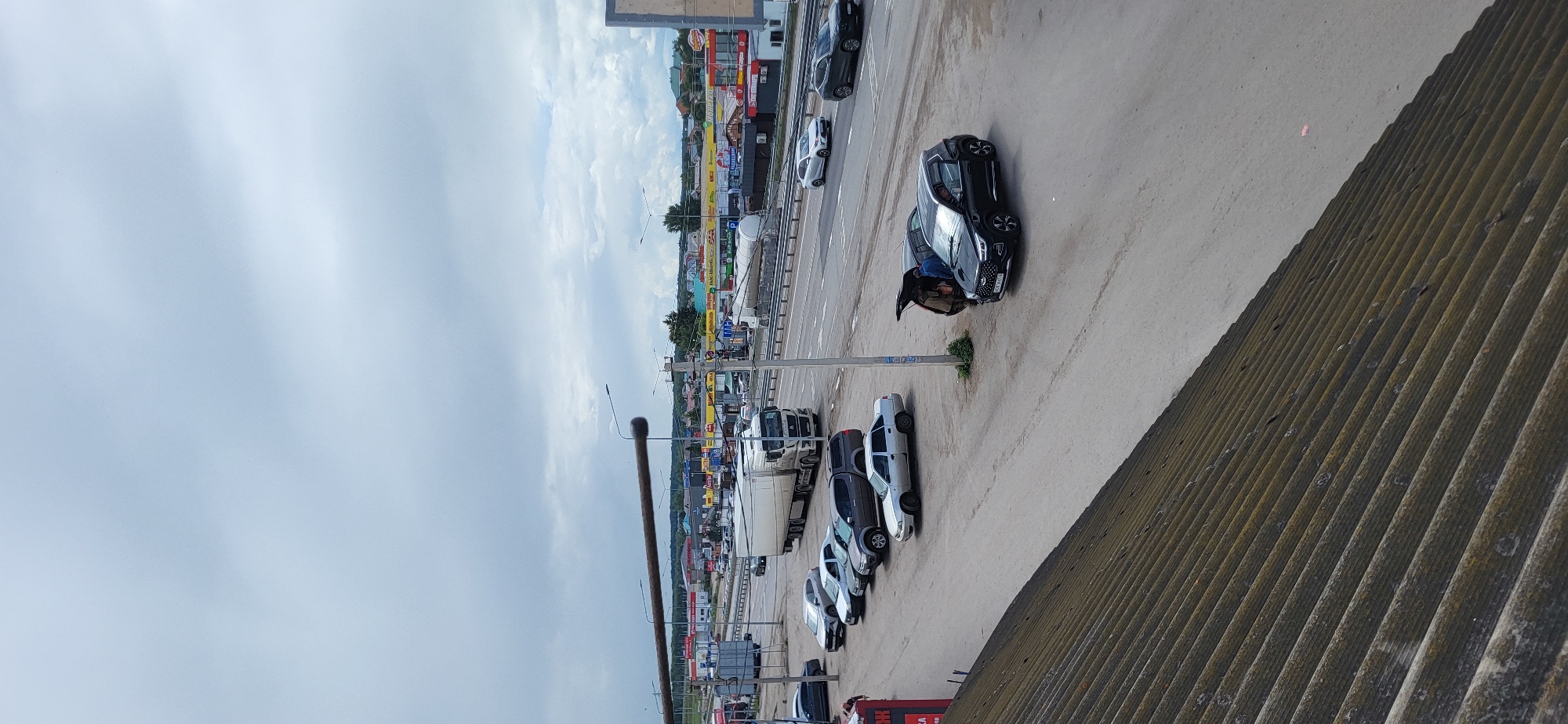 This image depicts a daytime scene in Bulgakovo, Russia, with a focus on a parking area adjacent to a highway. The sky is overcast with a light, grayish-blue hue.
In the foreground, a corrugated, weathered roof covers the lower left portion of the image. A gravel and asphalt parking area is visible, with several cars parked. A dark-colored SUV, possibly a Chery Tiggo, has its trunk open and two individuals are interacting with it. Next to this SUV is a lighter-colored sedan. Further back, a line of parked cars includes a silver sedan, a gray SUV, a white sedan, and a darker sedan.
The highway runs horizontally across the mid-ground of the image. Traffic is visible on the highway, including a large white semi-truck with a trailer. Other vehicles, including sedans and SUVs, are also on the highway.
In the background, a commercial area with various buildings and businesses is visible. Signs with Russian lettering are present on the buildings. The overall impression is one of a busy roadside commercial zone with a highway passing through.